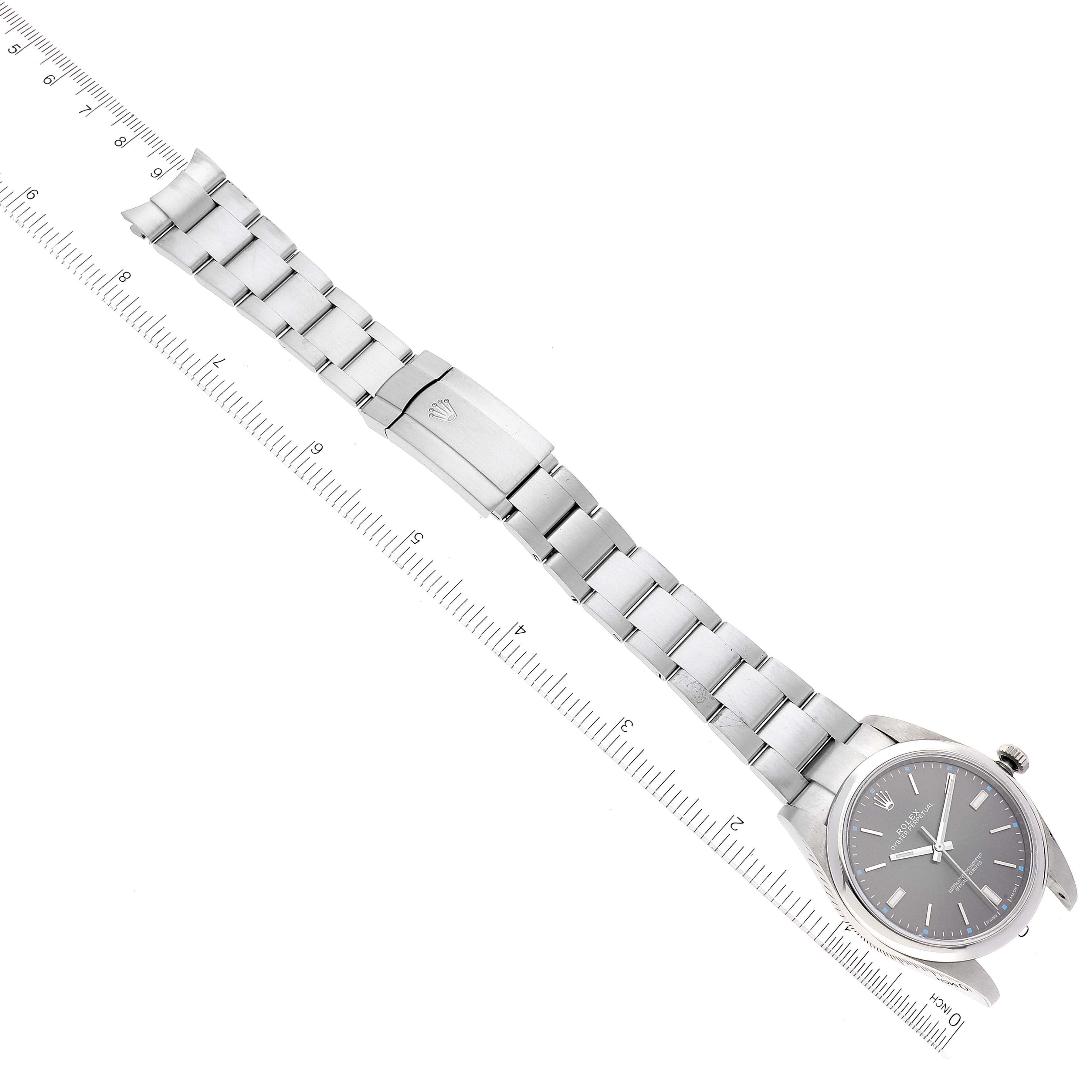 The image shows a Rolex Oyster Perpetual 114300 Men's Stainless Steel Grey Dial 114300 Men's Stainless Steel Grey Dial watch laid flat with its bracelet and clasp visible, viewed from above next to a ruler.