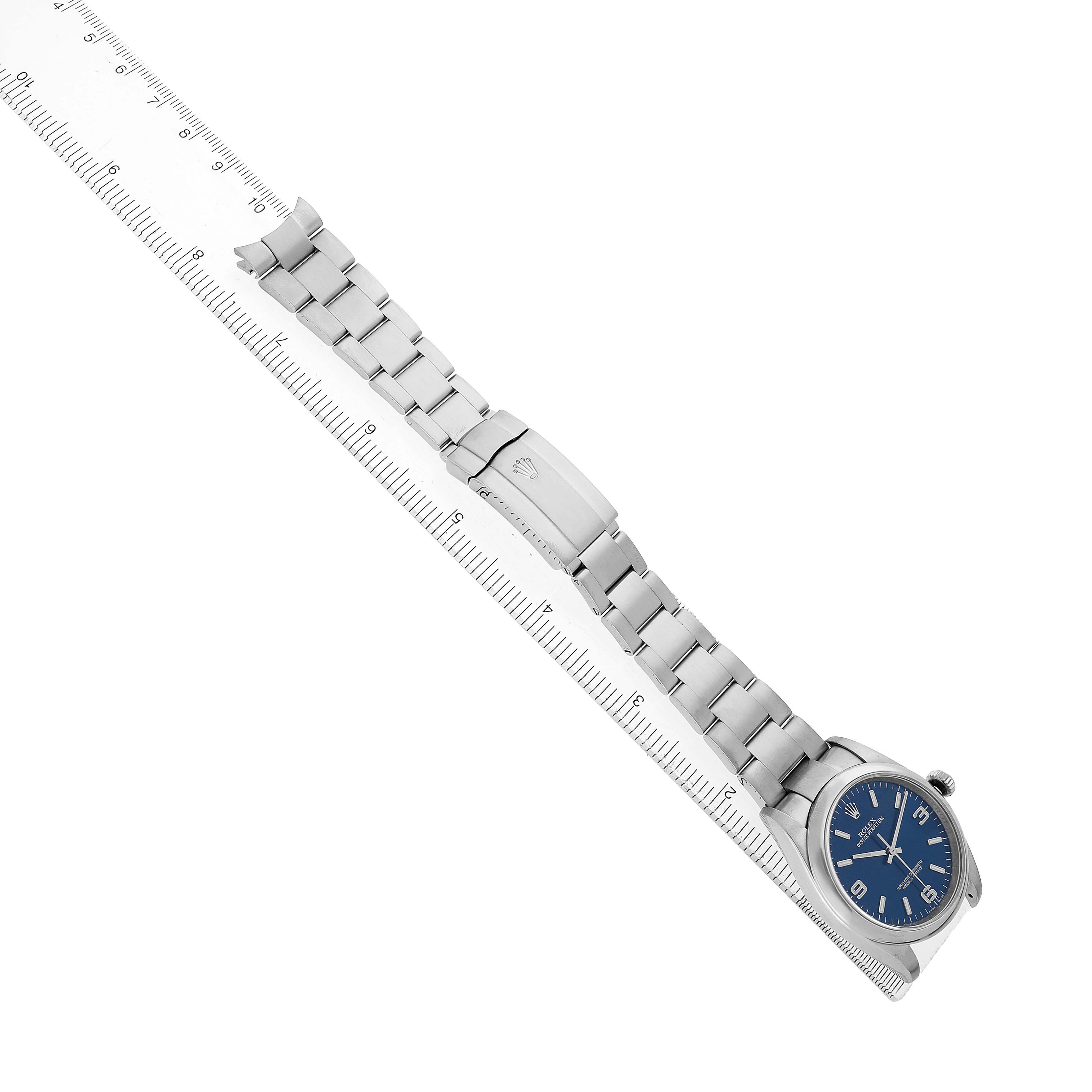 The Rolex Oyster Perpetual 116000 Men's Stainless Steel Blue Dial watch is shown at an angle displaying the face, bracelet, and clasp alongside a ruler.