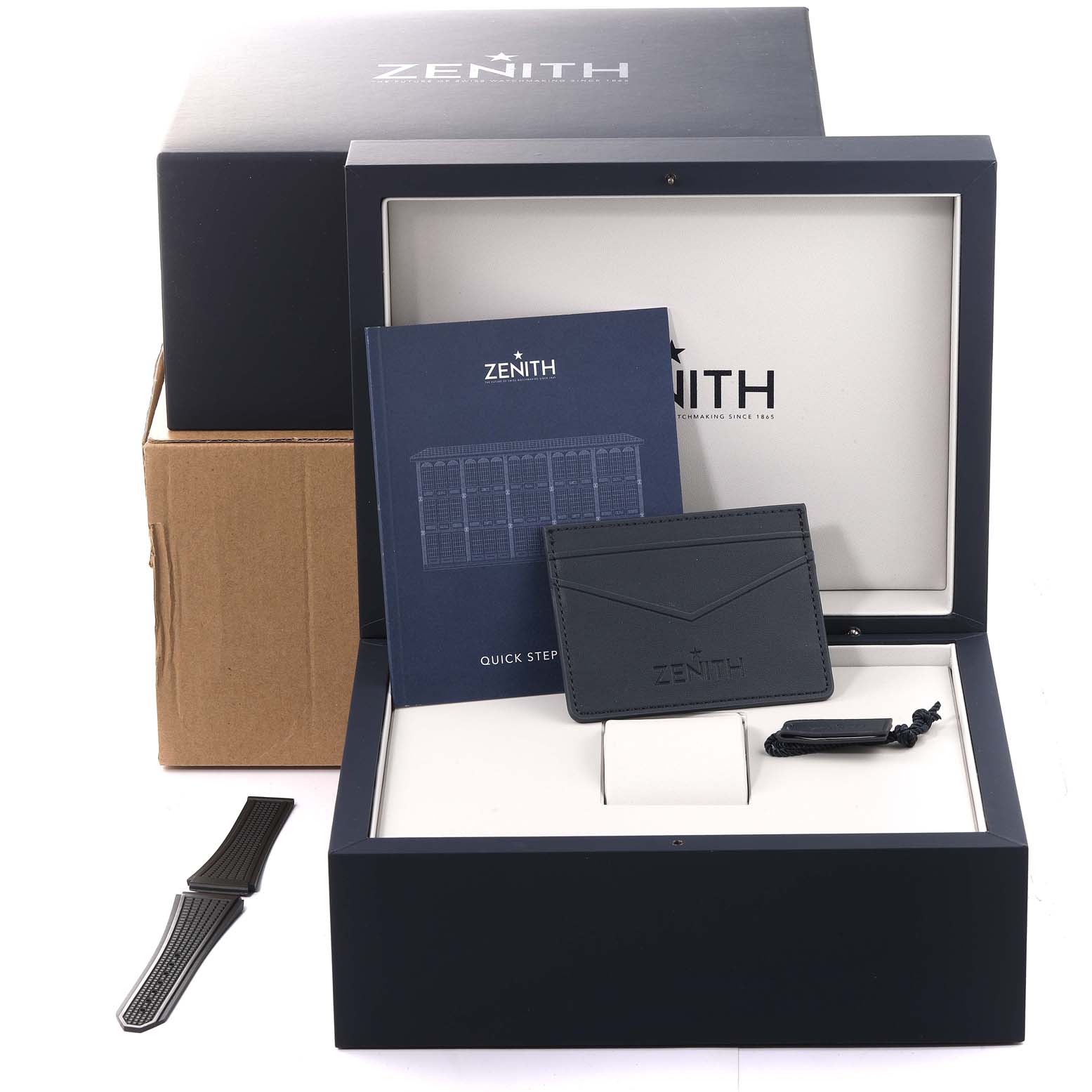 The image shows a Zenith Defy 95.9000.670/78.R782 Men's Titanium Grey Dial 95.9000.670/78.R782 Men's Titanium Grey Dial model watch packaging with booklets, an extra strap, and a card holder from a top-front angle.