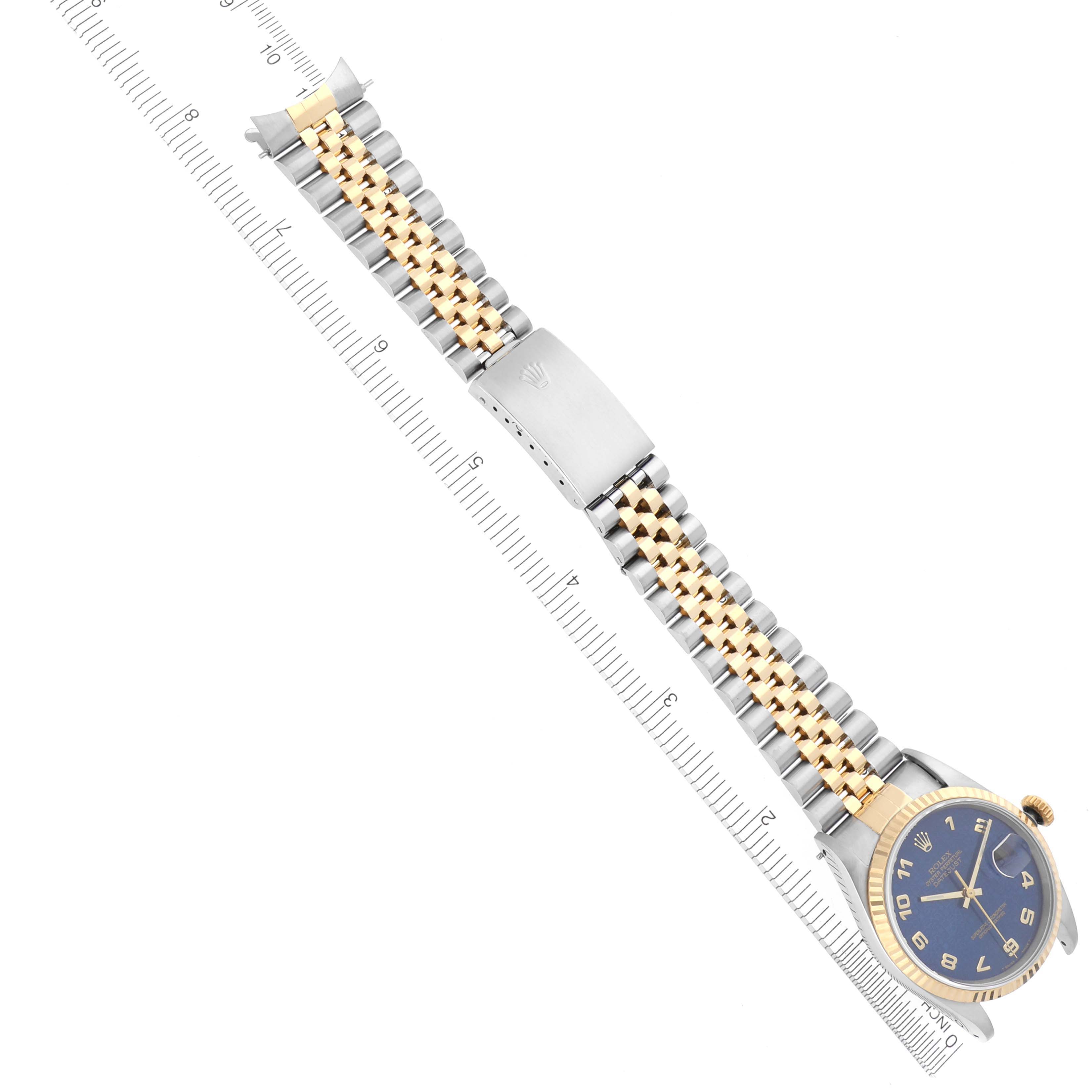 The Rolex Datejust 16233 Men's Steel and Gold (two tone) Blue Dial 16233 Men's Steel and Gold (two tone) Blue Dial watch is shown from a top-down angle, highlighting its blue dial, gold bezel, and Jubilee bracelet alongside a ruler.