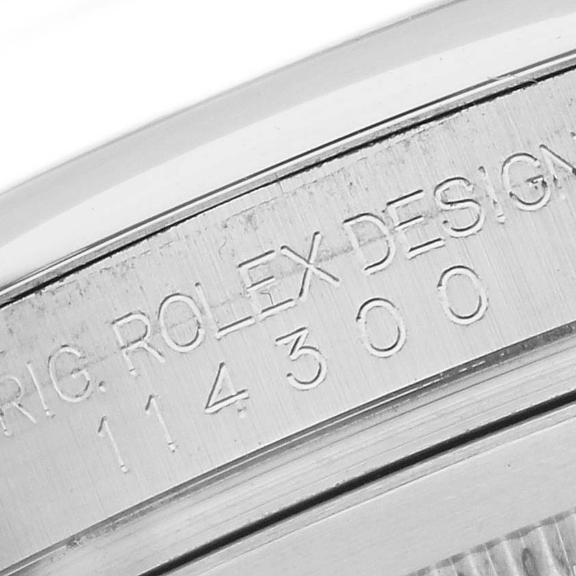 The image shows a close-up of the engraved edge of a Rolex Oyster Perpetual 114300 Men's Stainless Steel Grey Dial watch, highlighting the model number and branding.
