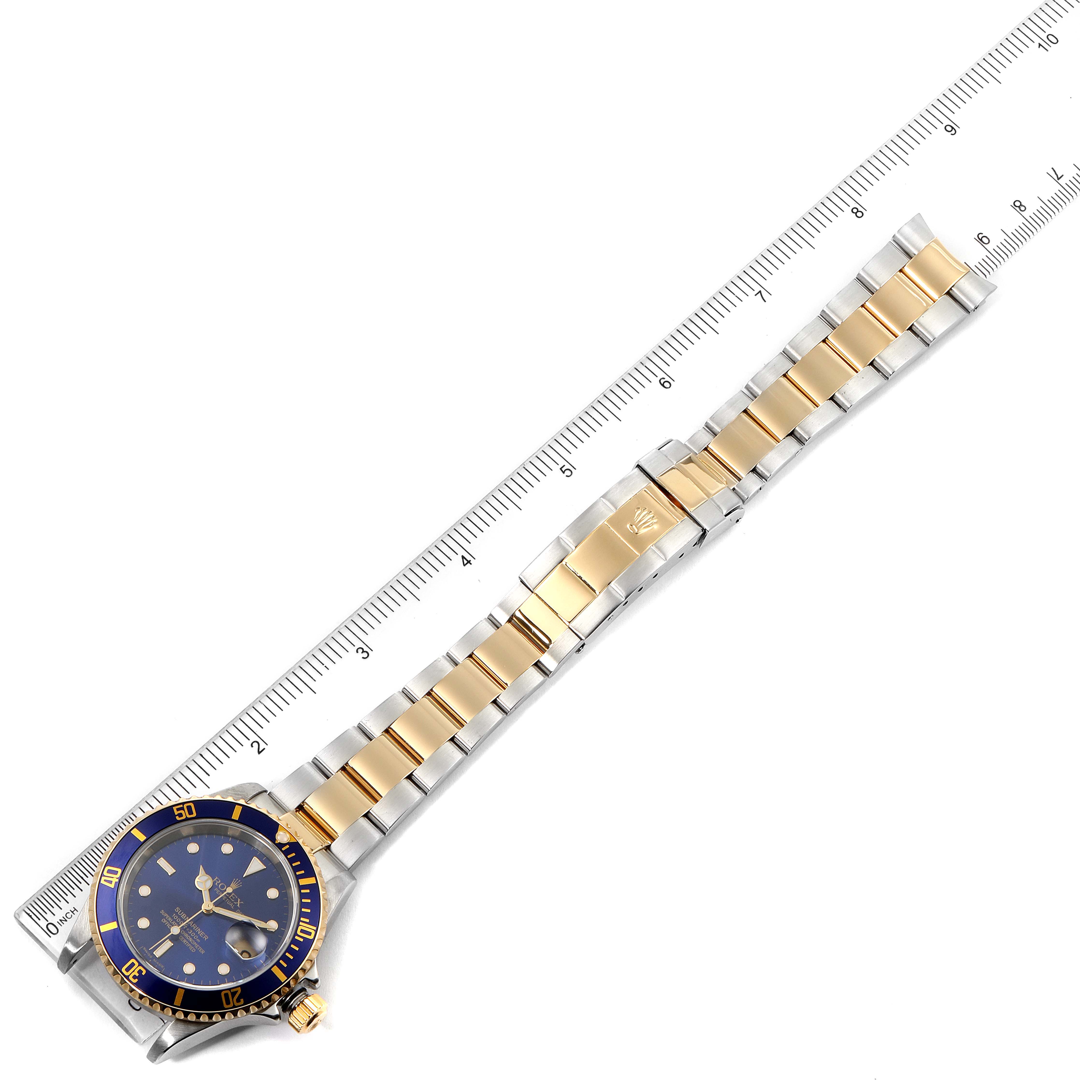 The image shows a Rolex Submariner 16613 Men's Steel and Gold (two tone) Blue Dial 16613 Men's Steel and Gold (two tone) Blue Dial watch laid flat with a full view of its band, resting next to a ruler.