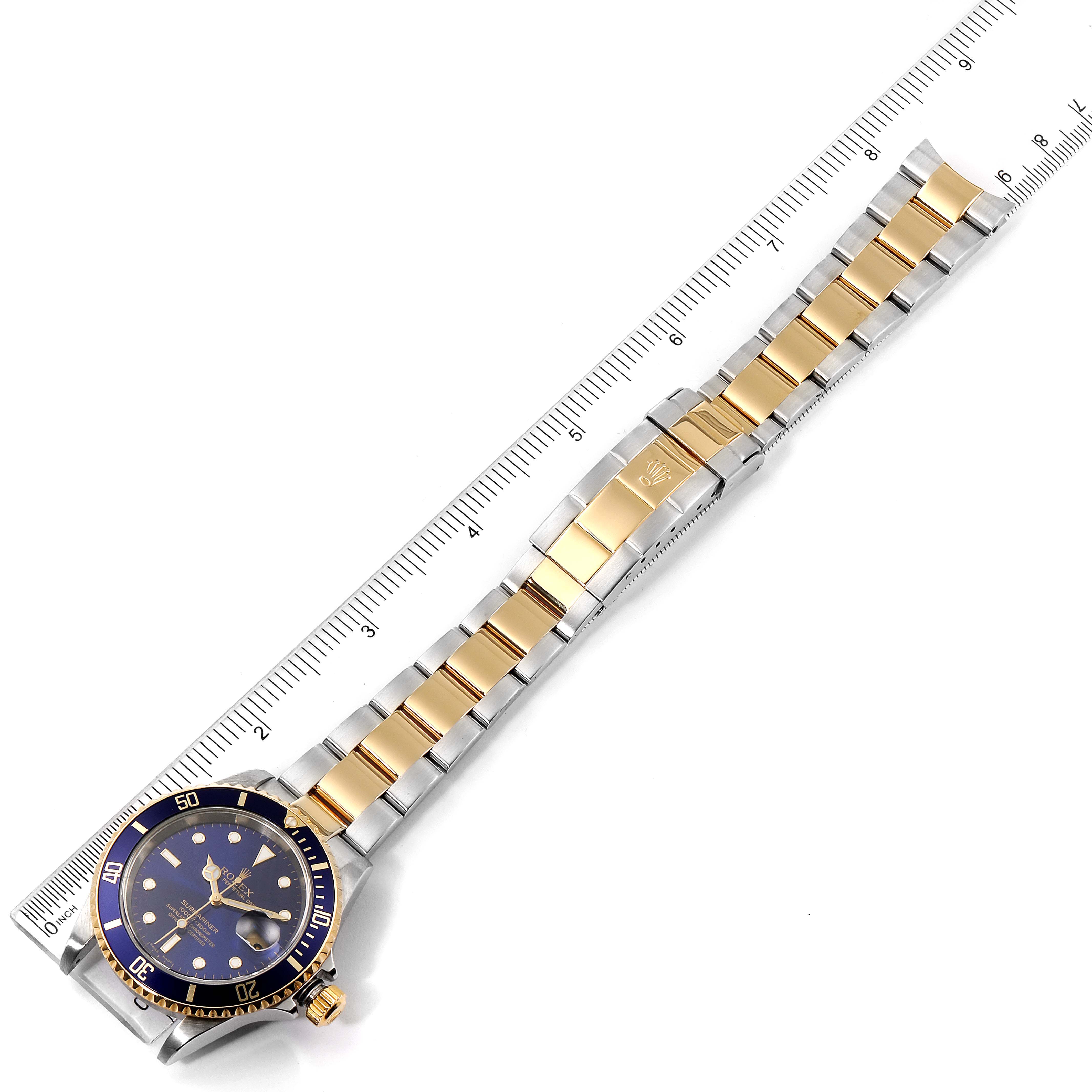 The image shows a Rolex Submariner 16613 Men's Steel and Gold (two tone) Blue Dial 16613 Men's Steel and Gold (two tone) Blue Dial watch laid flat, highlighting its gold and silver bracelet, bezel, dial, and clasp, with a ruler for scale.