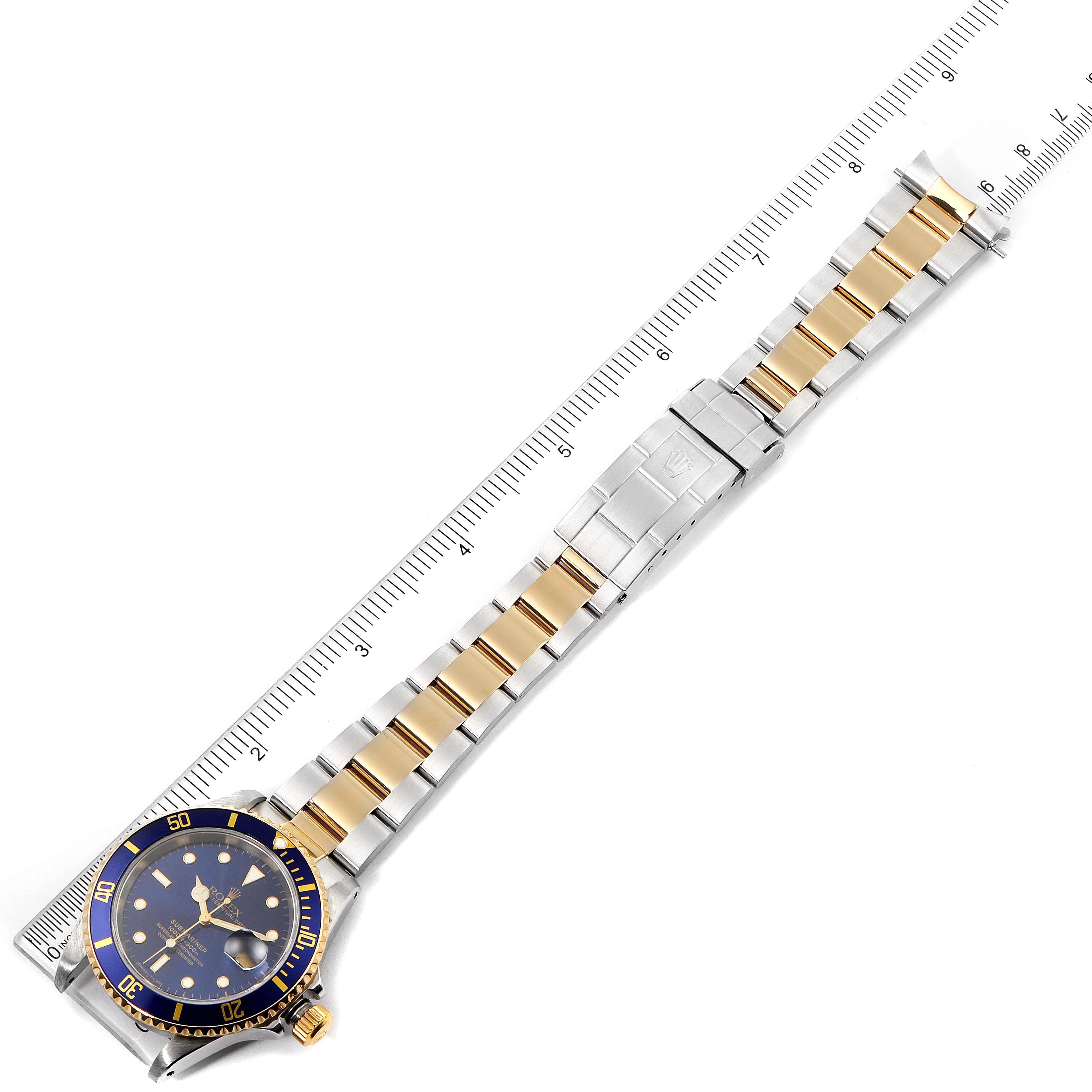 The image shows a Rolex Submariner 16613 Men's Steel and Gold (two tone) Blue Dial 16613 Men's Steel and Gold (two tone) Blue Dial watch laid flat with a ruler next to it, highlighting its band and face.