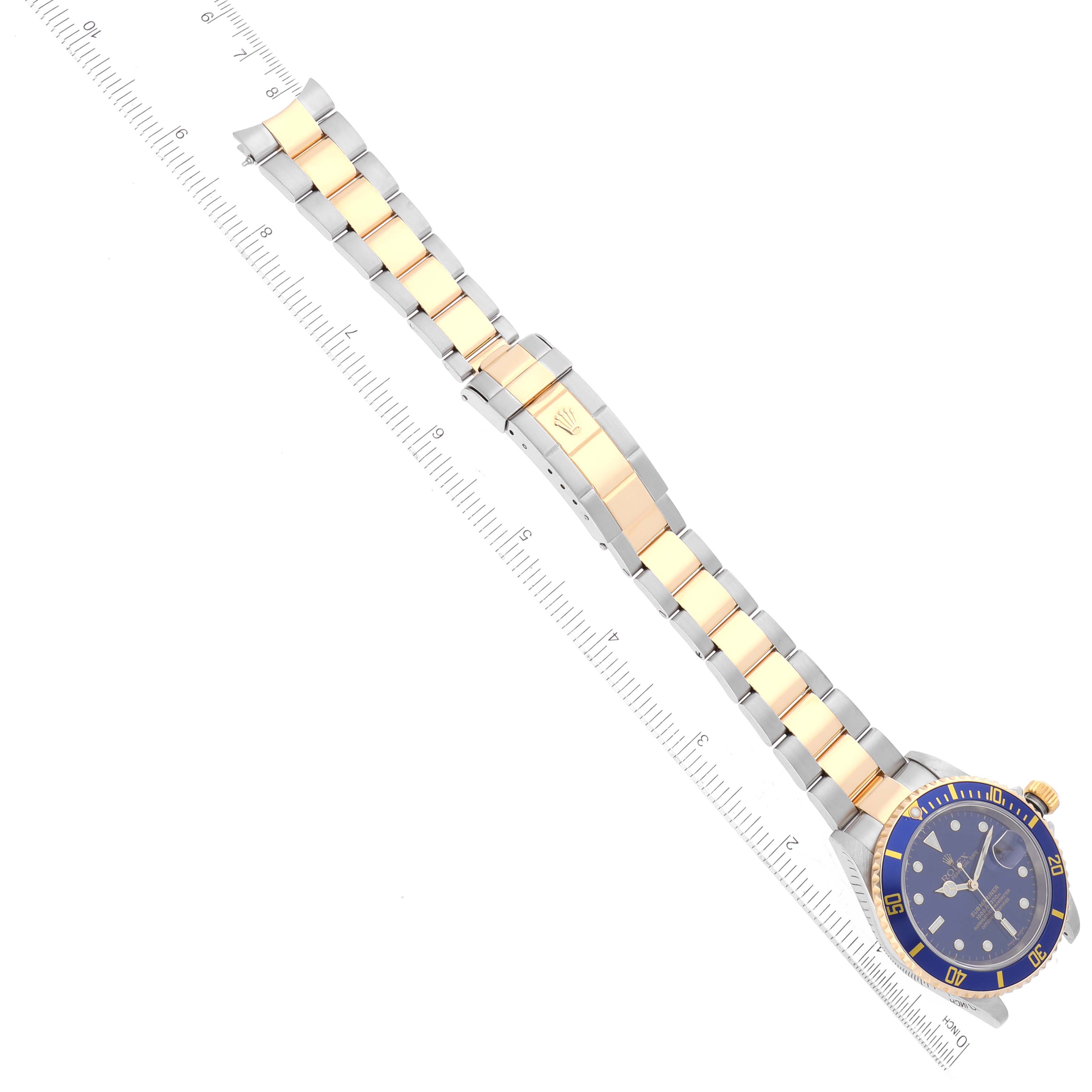 The image shows a top-down view of a Rolex Submariner 16613 Men's Steel and Gold (two tone) Blue Dial 16613 Men's Steel and Gold (two tone) Blue Dial watch, featuring its blue dial, bezel, and two-tone metal bracelet, alongside a ruler for scale.