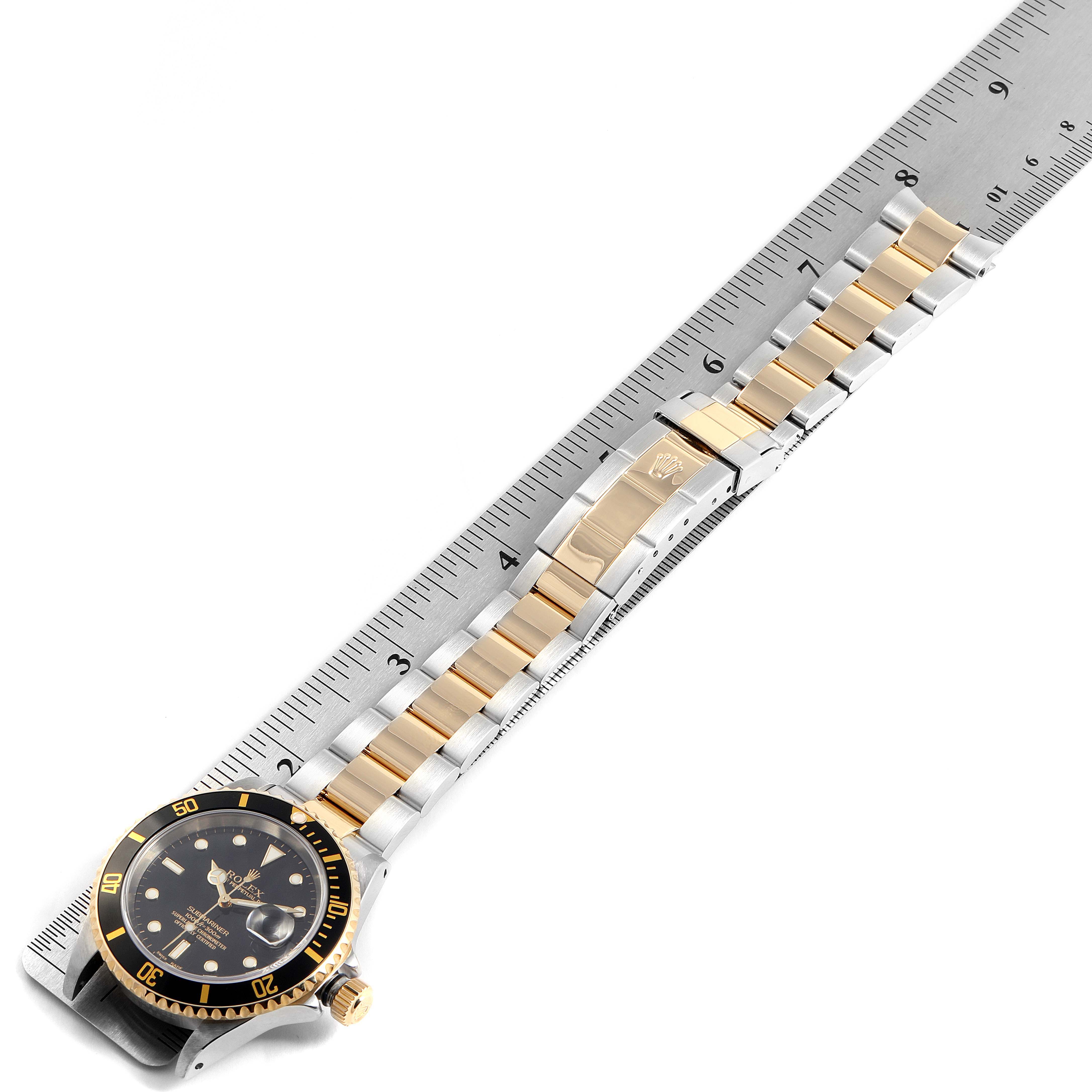 The image shows a top-down view of a Rolex Submariner 16613 Men's Steel and Gold (two tone) Black Dial 16613 Men's Steel and Gold (two tone) Black Dial watch, including its bracelet, bezel, and clasp, next to a ruler.