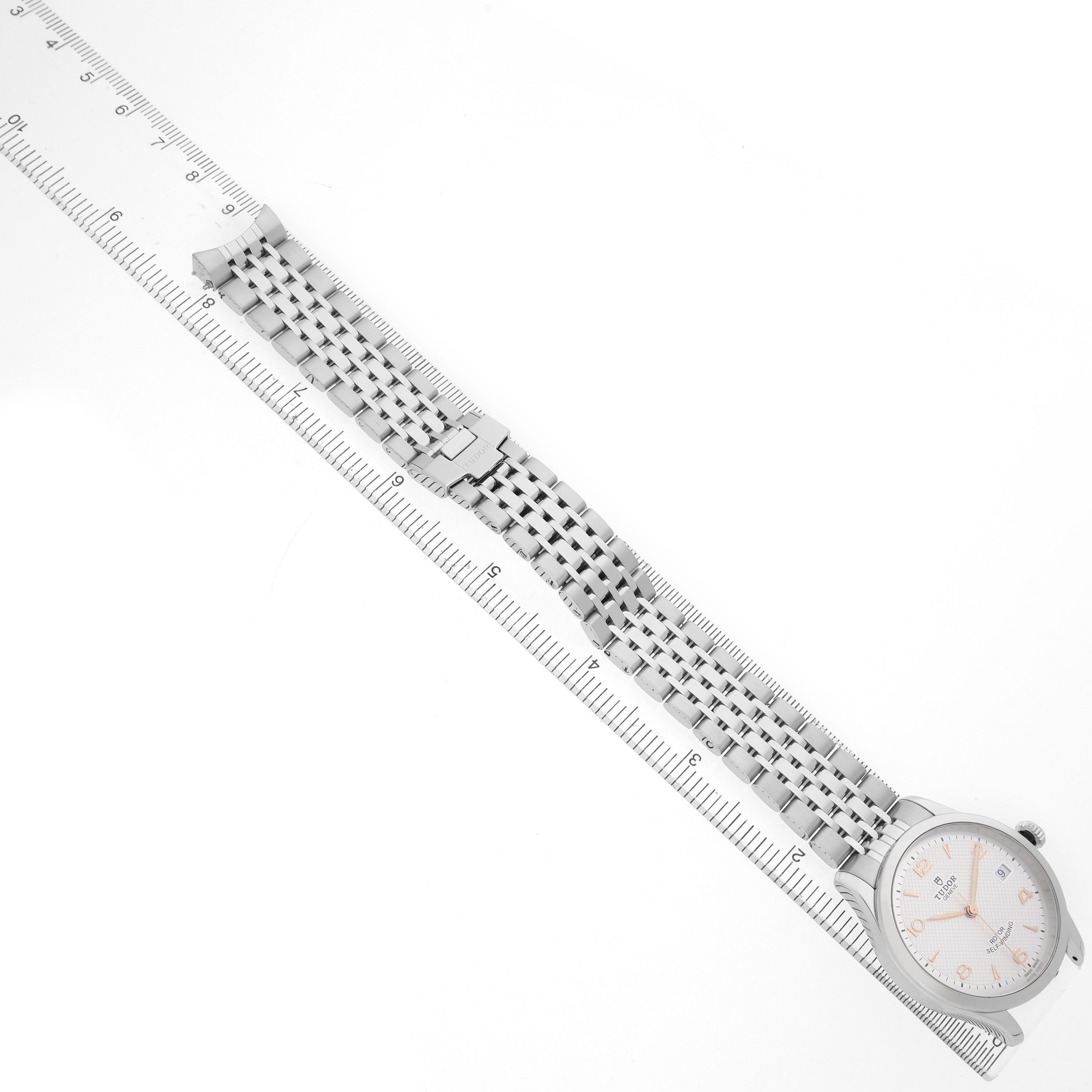 The Tudor 1926 M91450-0001 Men's Stainless Steel Silver Dial watch is shown from a top view, displaying its face, strap, and clasp alongside a ruler for scale.