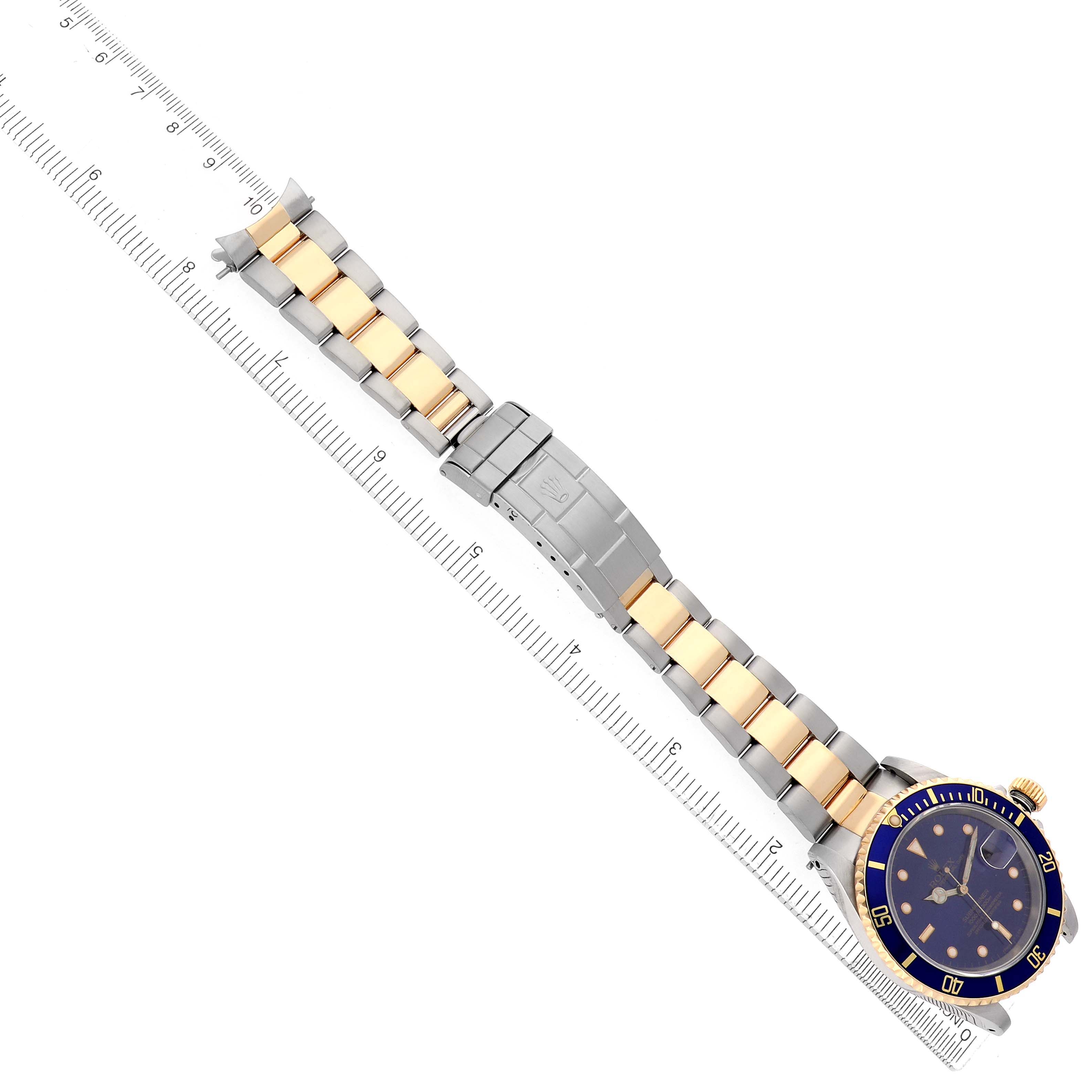 The image shows a Rolex Submariner 16613 Men
s Steel and Gold (two tone) Blue Dial watch from an overhead angle, displaying the full bracelet, clasp, and watch face next to a ruler.