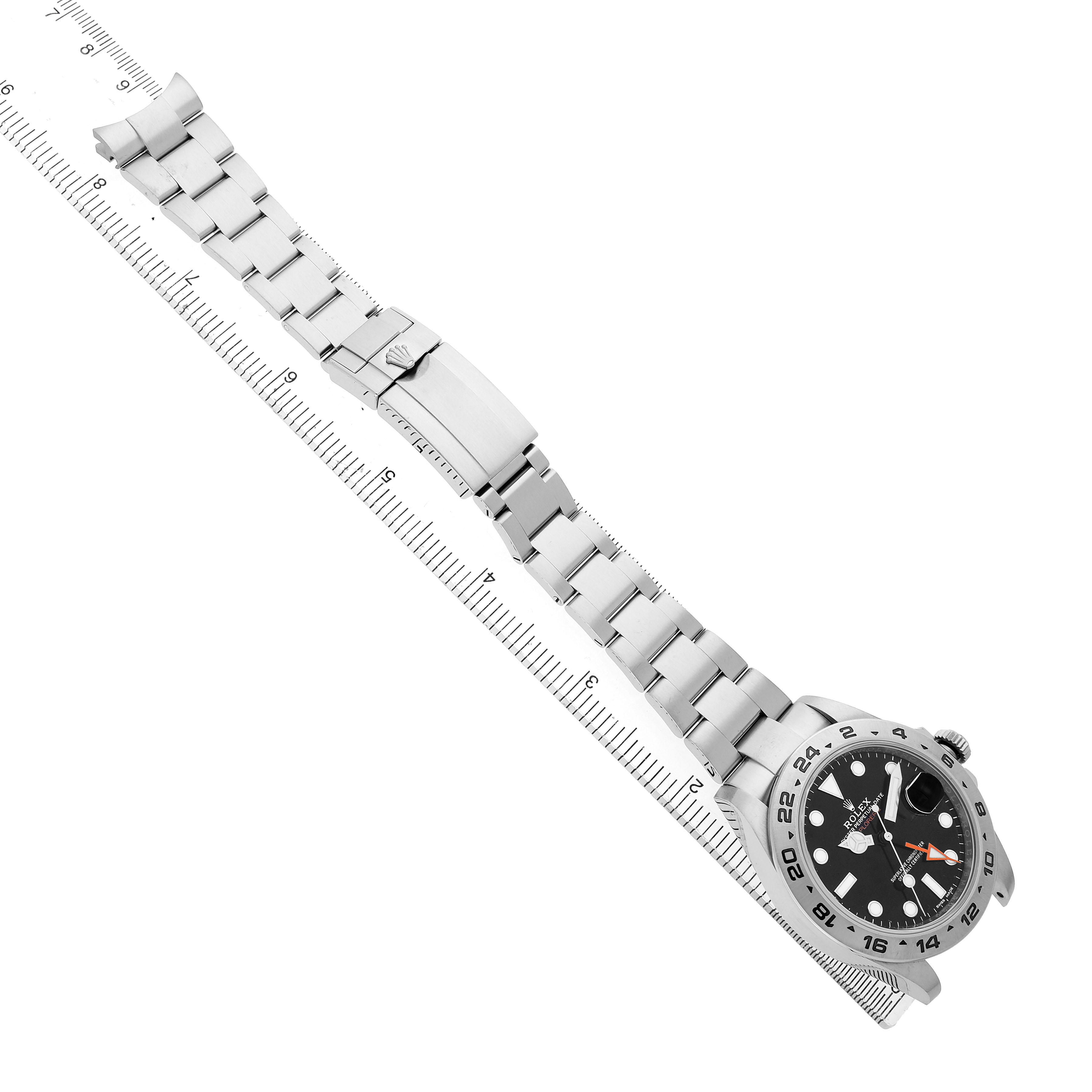 The Rolex Explorer 216570 Men's Stainless Steel Black Dial watch is shown at a diagonal angle, displaying the full bracelet, clasp, and watch face against a ruler backdrop.