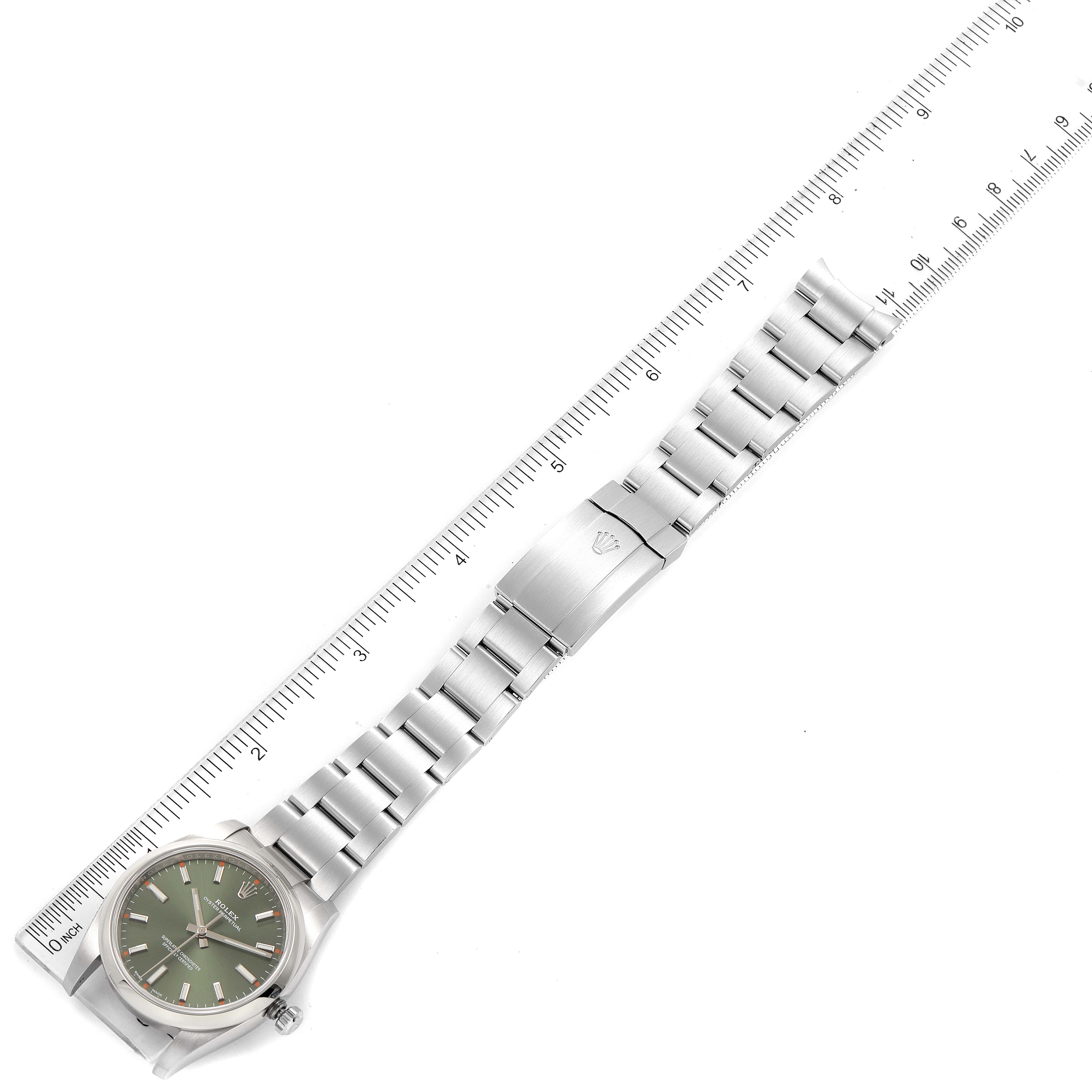 The image shows a Rolex Oyster Perpetual 114200 Men
s Stainless Steel Green Dial 114200 Men
s Stainless Steel Green Dial watch laid flat next to a ruler, displaying its face, dial, bracelet, and clasp.
