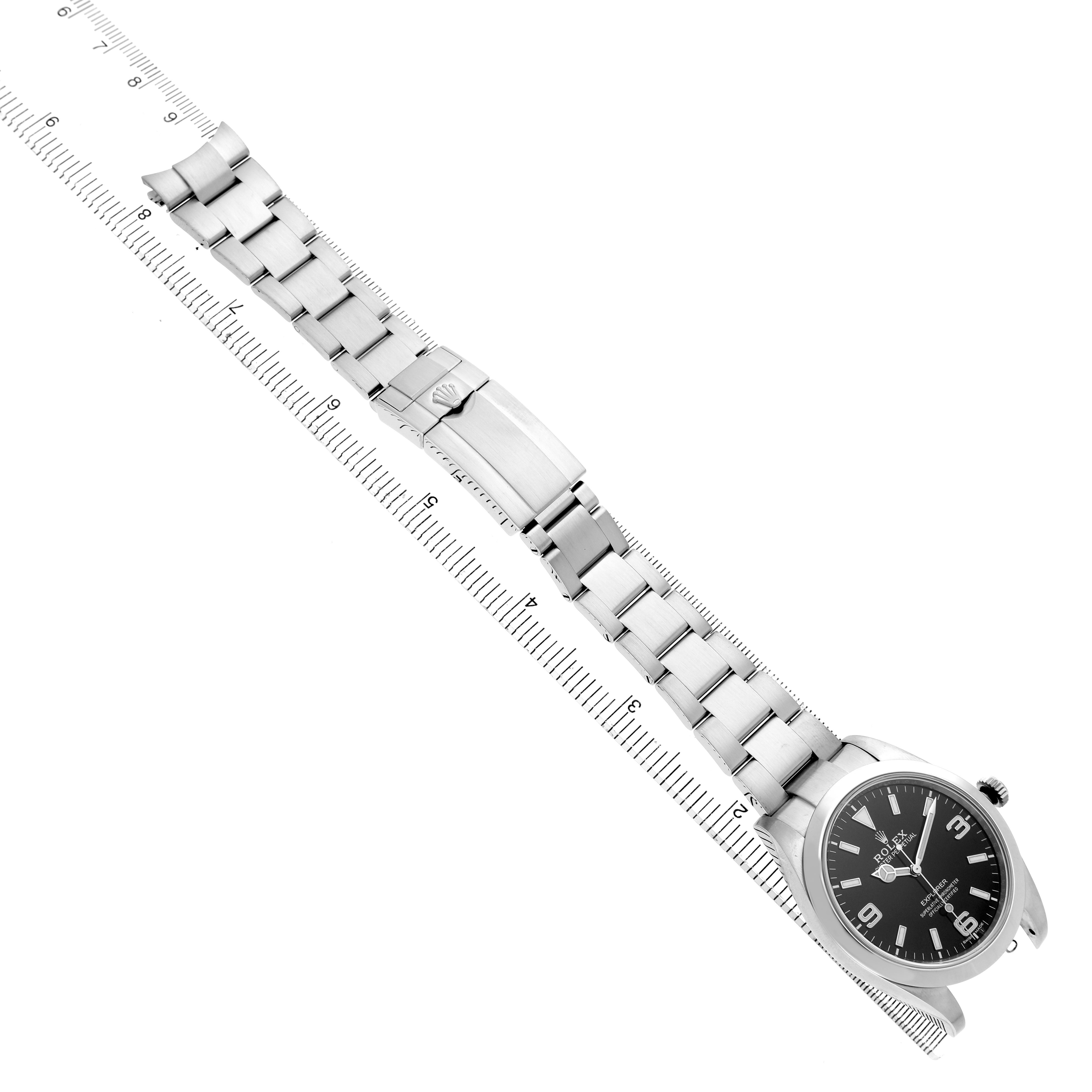 The Rolex Explorer 214270 Men's Stainless Steel Black Dial watch is displayed at an angle, showcasing the face, bracelet, and clasp alongside a measuring ruler.