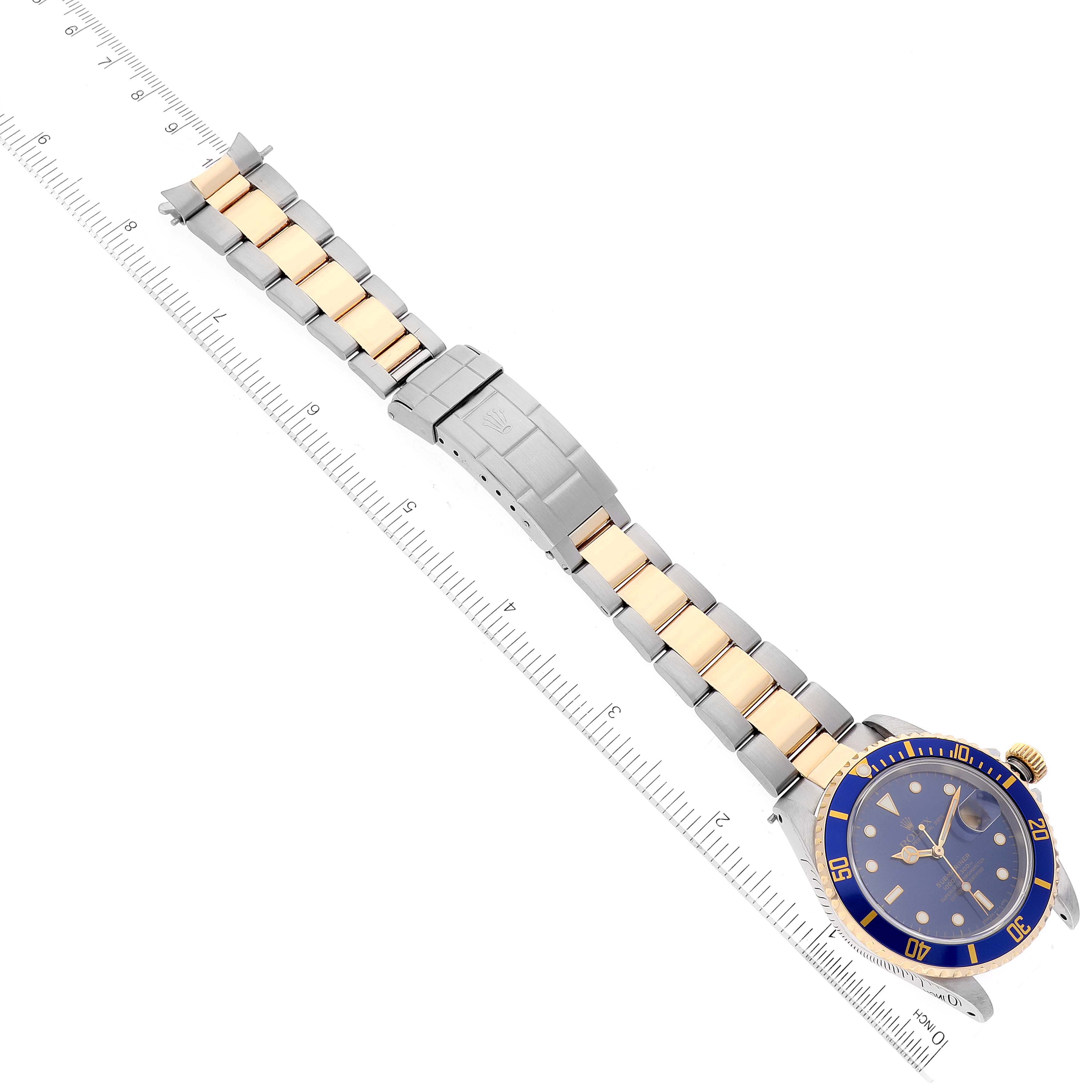 The image shows a Rolex Submariner 16613 Men
s Steel and Gold (two tone) Blue Dial 16613 Men
s Steel and Gold (two tone) Blue Dial watch with a blue dial and two-tone bracelet, viewed from a top-down angle alongside a ruler.