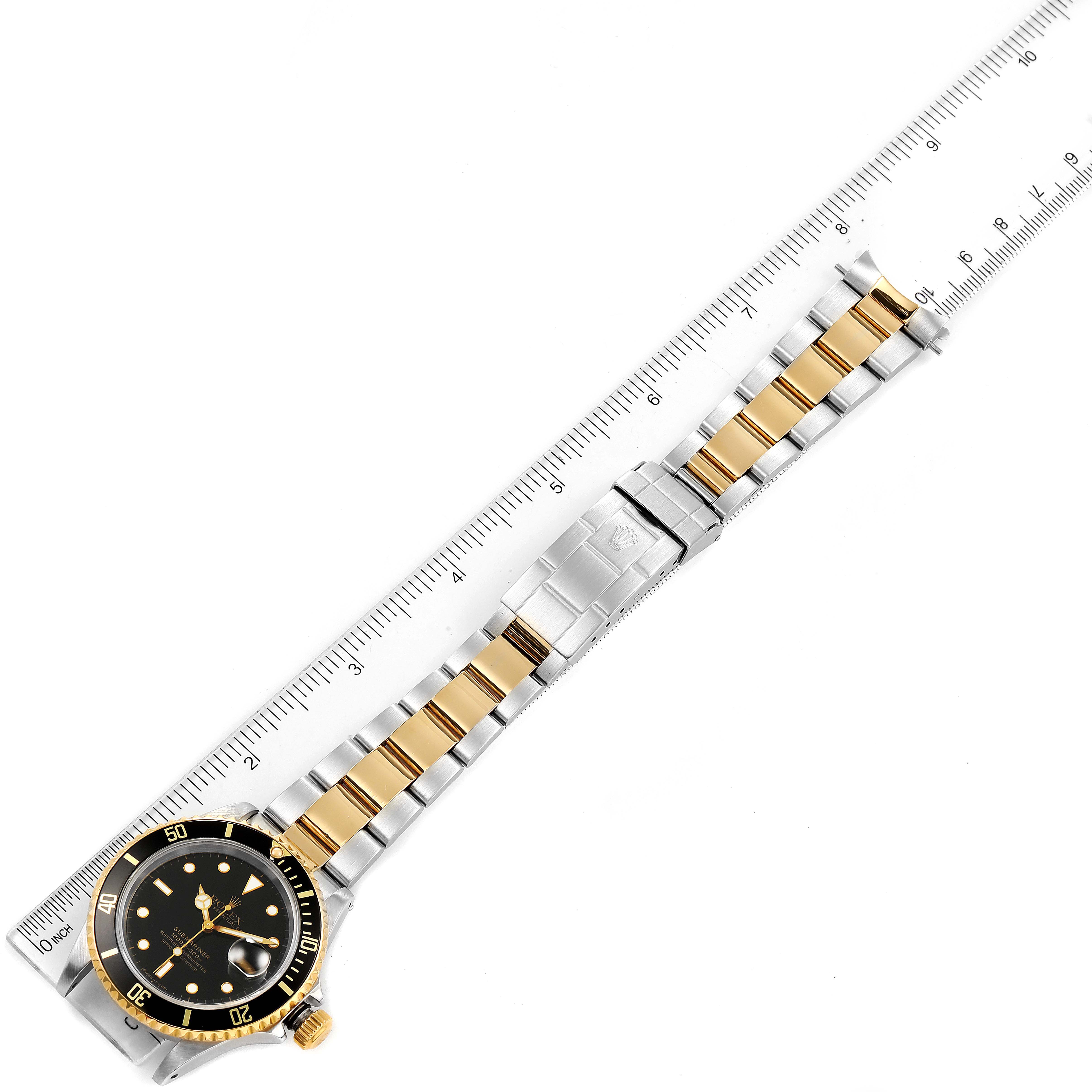 The Rolex Submariner 16613 Men's Steel and Gold (two tone) Black Dial 16613 Men's Steel and Gold (two tone) Black Dial watch is shown from an aerial view, displaying the whole watch, including the bezel, dial, and bracelet next to a ruler.