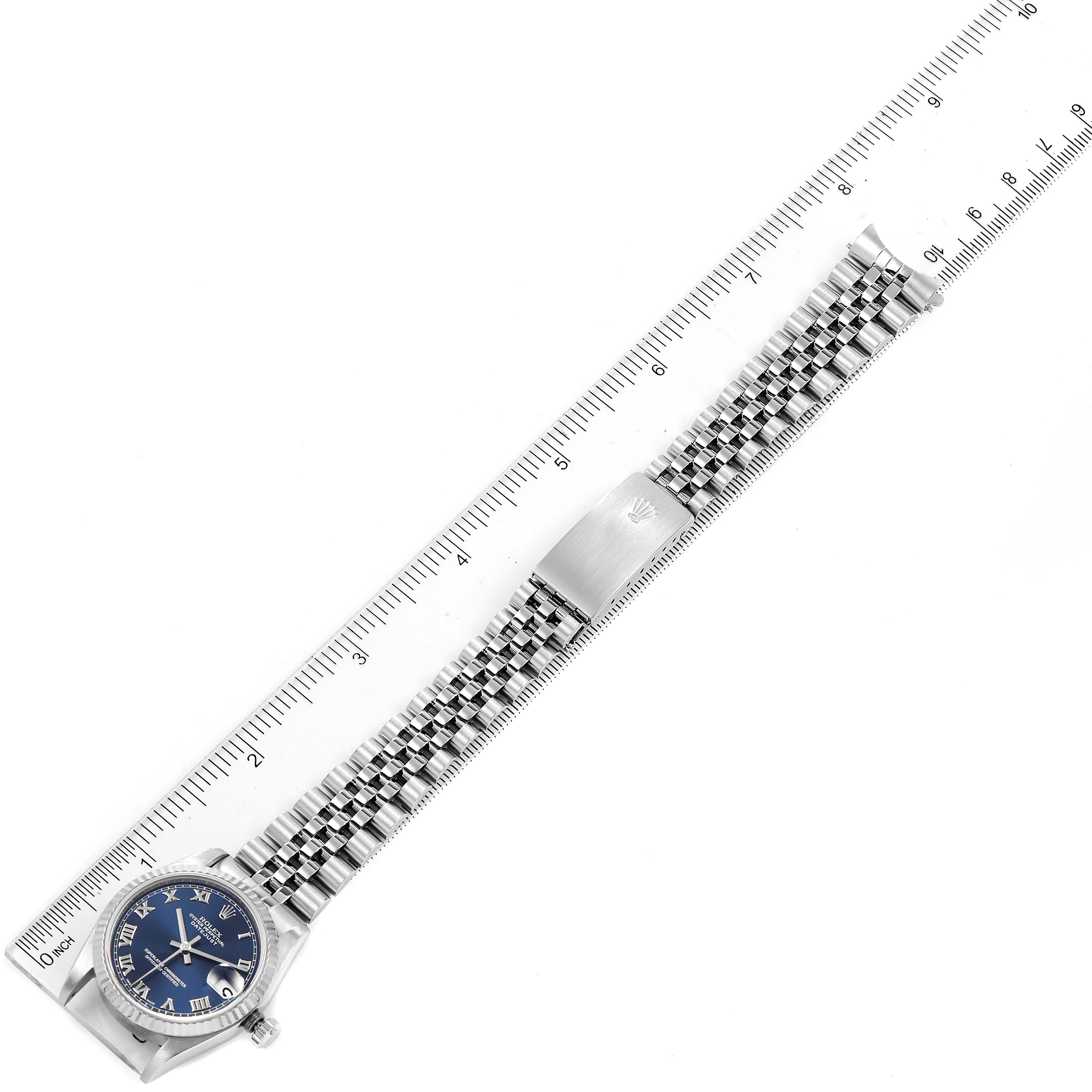 The Mid-Size Rolex watch is shown from a top-down angle, displaying its blue dial, bezel, and full bracelet beside a ruler for scale.