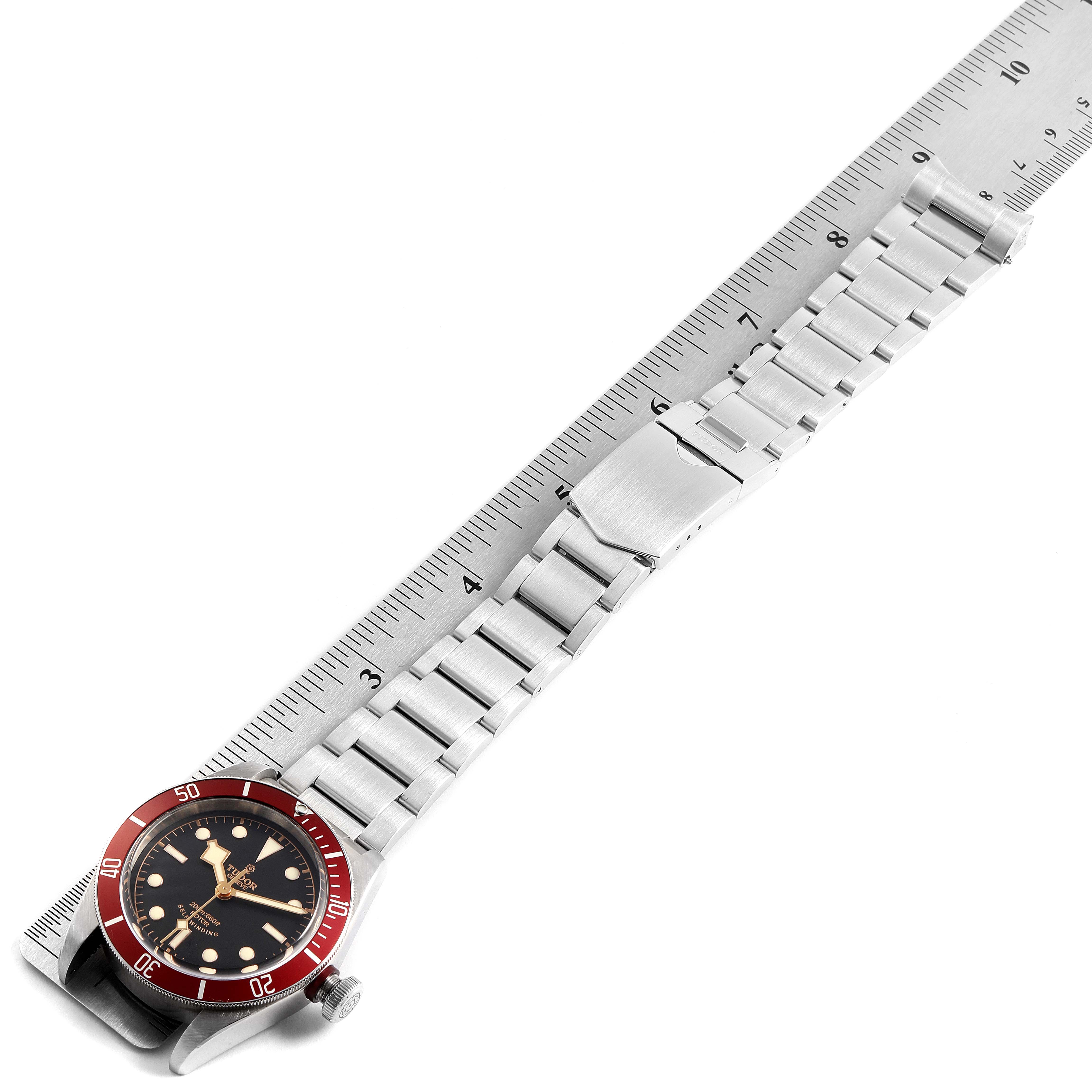 The image shows the Tudor Heritage Black Bay 79220R Men's Stainless Steel Black Dial 79220R Men's Stainless Steel Black Dial watch with a red bezel and metal bracelet, placed next to a ruler.