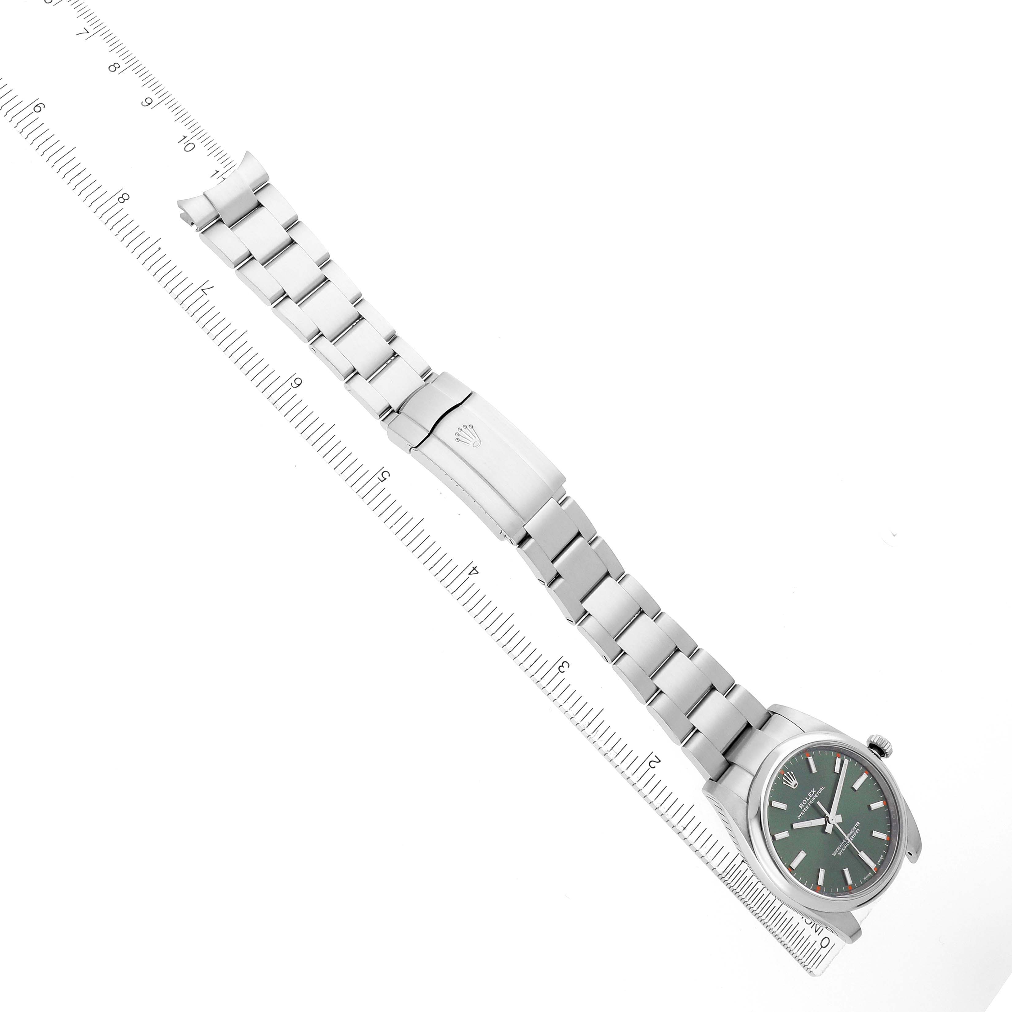 The Rolex Oyster Perpetual 114200 Men's Stainless Steel Green Dial watch is displayed fully extended with a ruler alongside, showcasing the face, bracelet, and clasp.