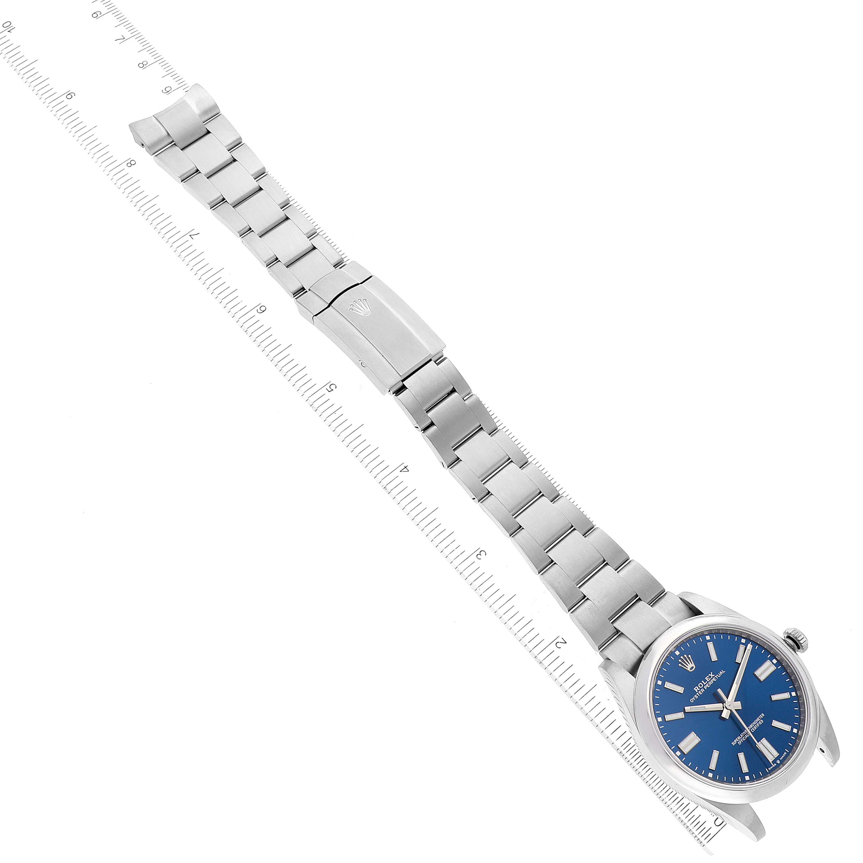 The image shows a top view of a Rolex Oyster Perpetual 124300 Men
s Stainless Steel Blue Dial 124300 Men
s Stainless Steel Blue Dial watch with a blue dial and metal bracelet laid next to a ruler.