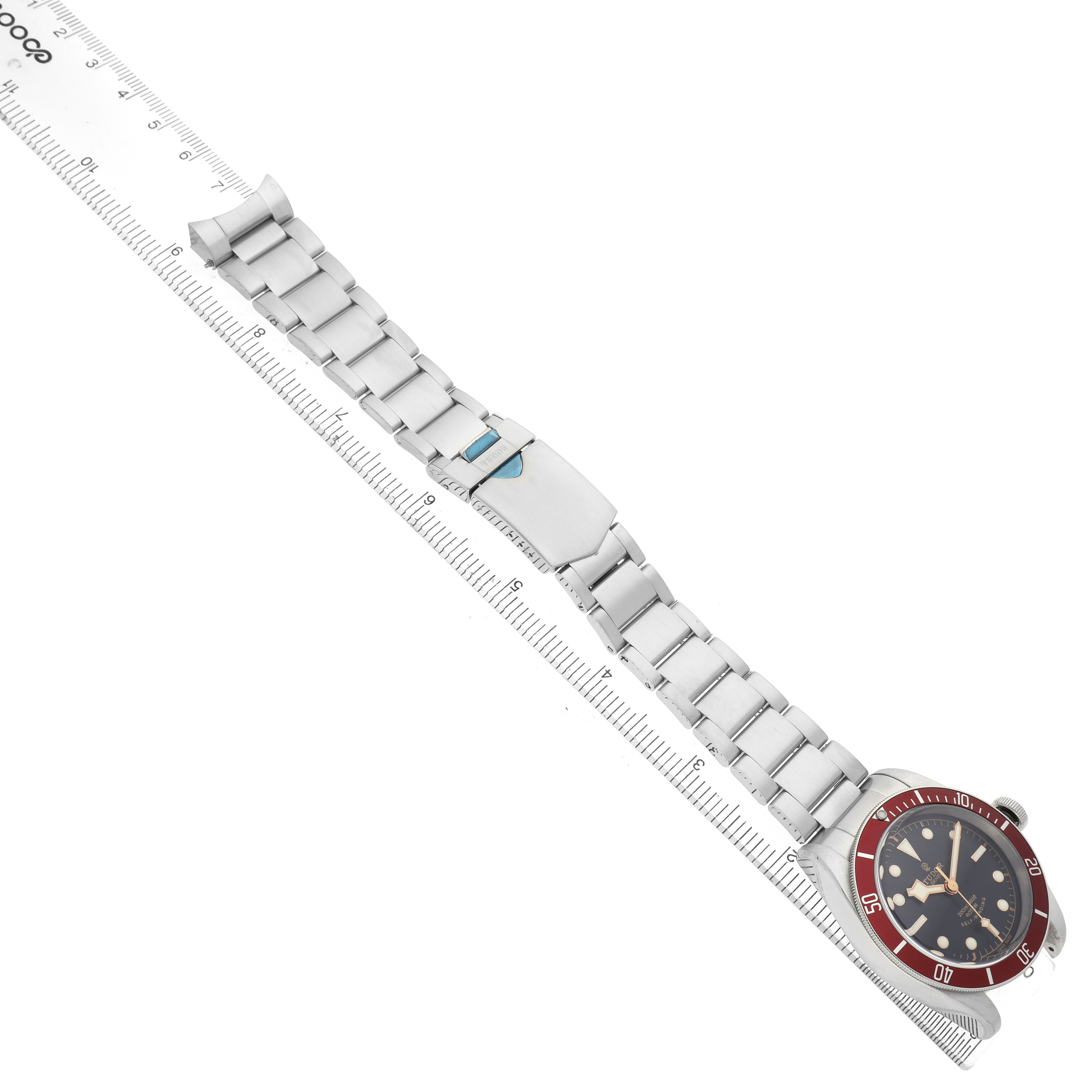 The Tudor Heritage Black Bay 79220R Men's Stainless Steel Black Dial watch is shown flat with the bracelet fully extended, lying diagonally with a ruler alongside.
