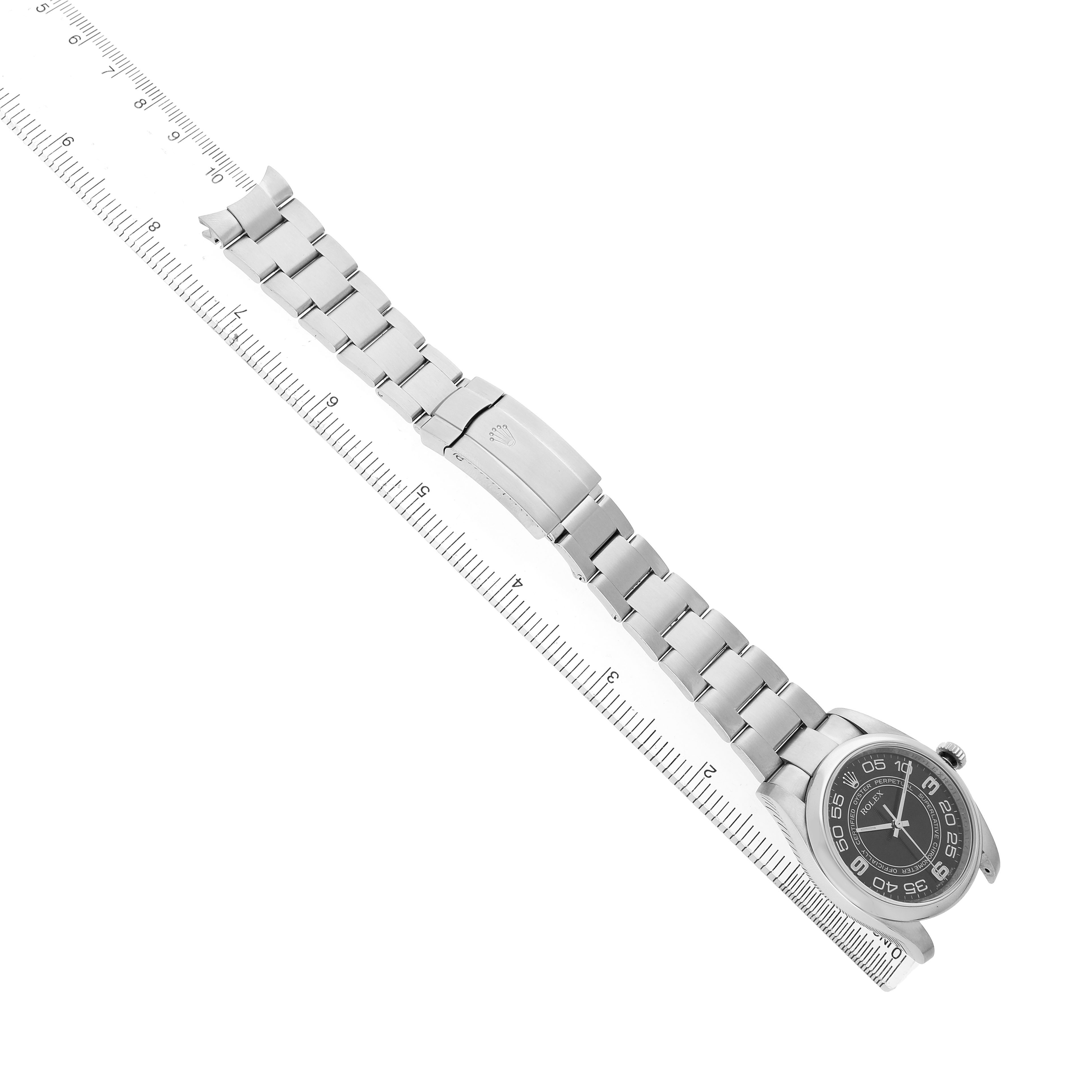 The Rolex Oyster Perpetual 116000 Men's Stainless Steel Black Dial watch is shown from an overhead angle, displaying the full bracelet and face alongside a ruler for scale.