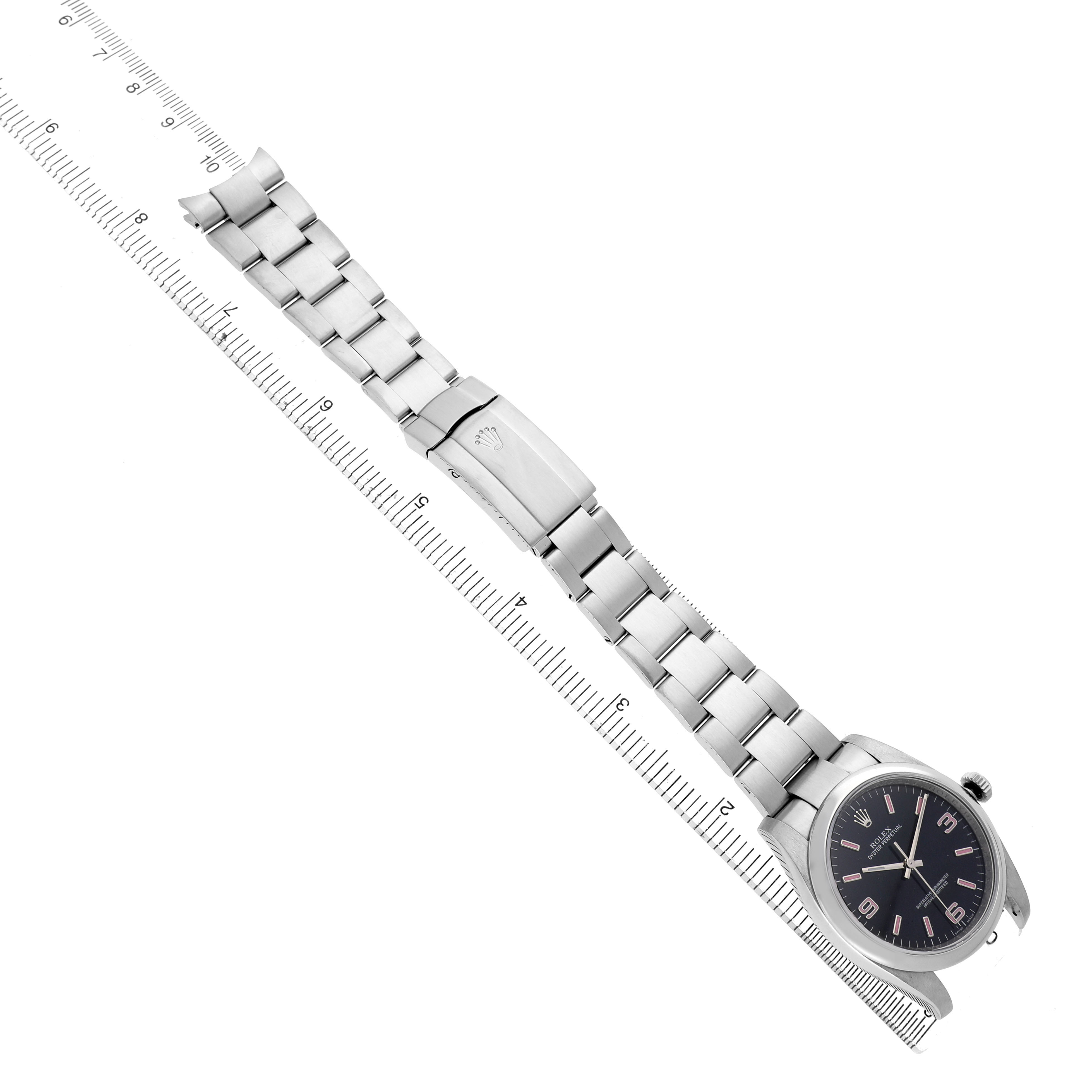 The Rolex Oyster Perpetual 116000 Men's Stainless Steel Black Dial watch is shown at an oblique angle, displaying the full bracelet and clasp on a ruler.