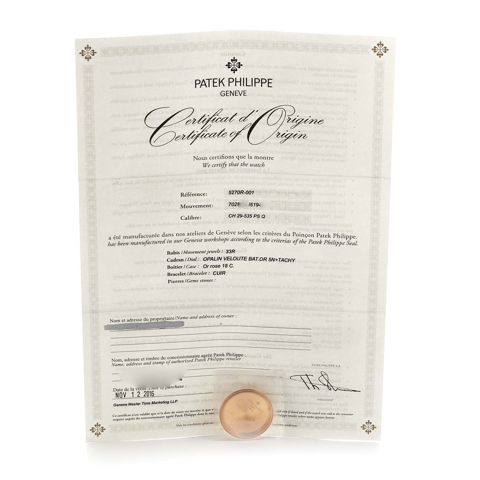 The image shows a Patek Philippe Complications 5270R-001 Men's Rose Gold Silver Dial model certificate of origin and a small accessory, likely a seal, from a slightly elevated angle.