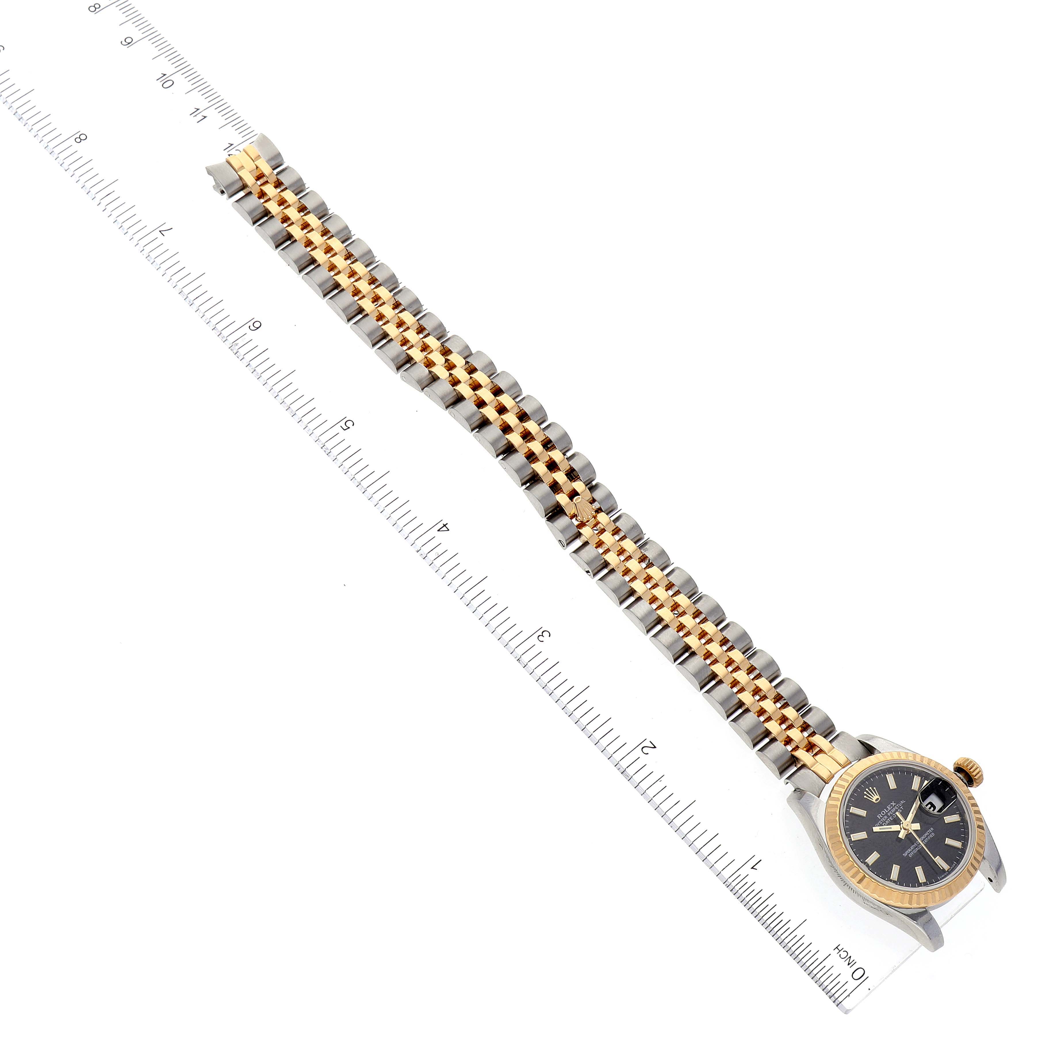 The image shows a top view of a Rolex Datejust 179173 Women's Steel and Gold (two tone) Black Dial 179173 Women's Steel and Gold (two tone) Black Dial watch with a gold and silver bracelet next to a ruler for scale.