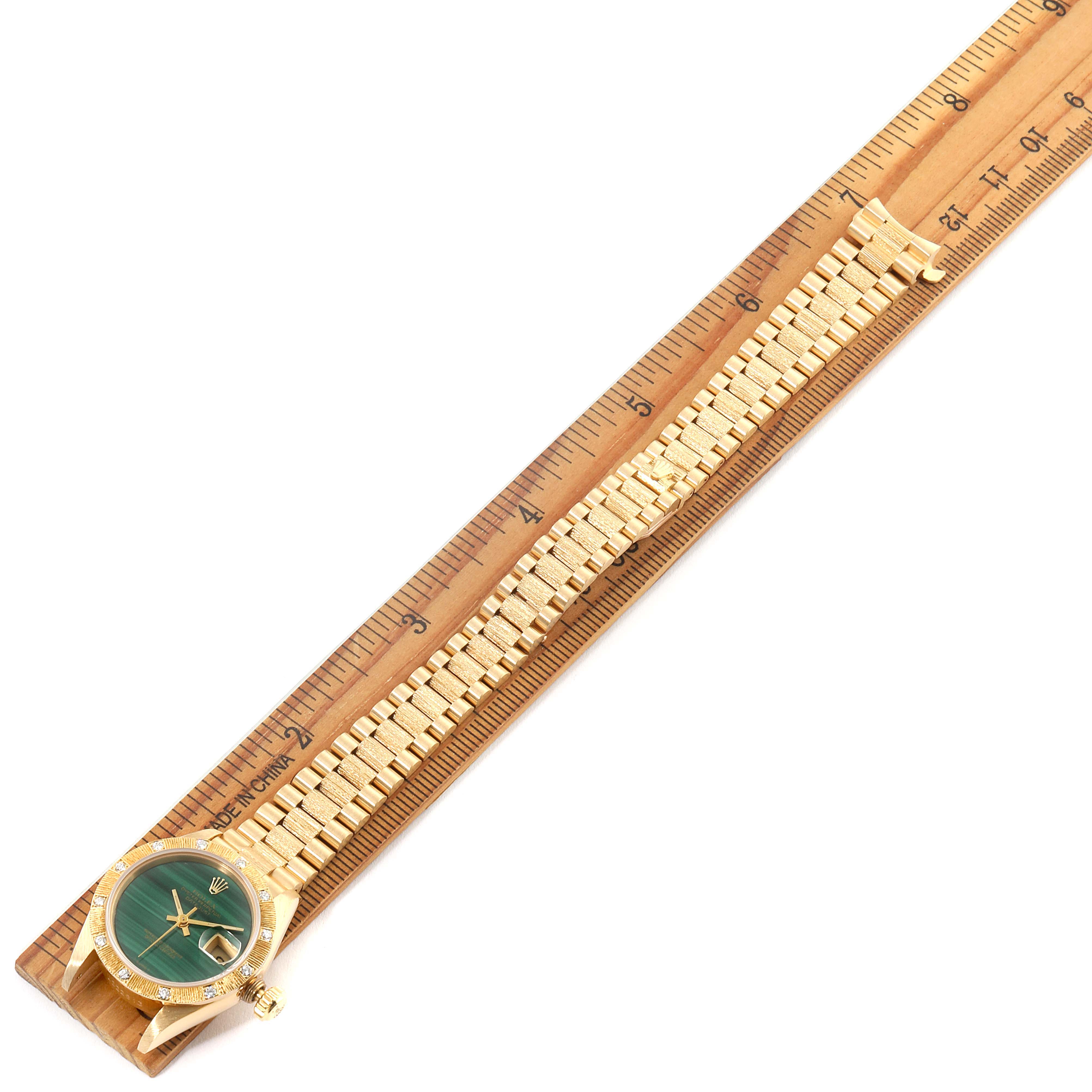 The President model Rolex watch is displayed diagonally on a ruler, showing the gold bracelet, green dial, and bezel.