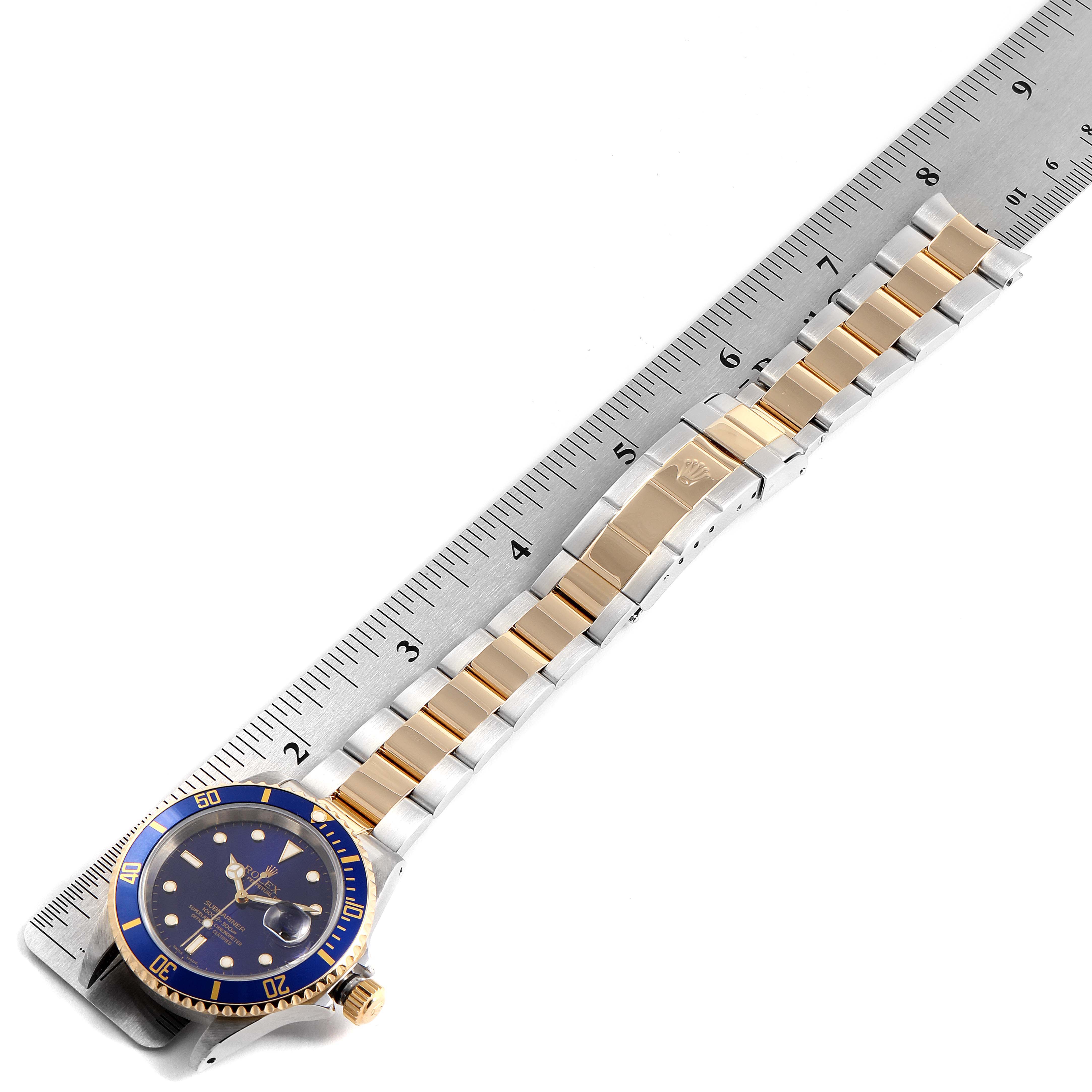The image shows a Rolex Submariner 16613 Men
s Steel and Gold (two tone) Blue Dial 16613 Men
s Steel and Gold (two tone) Blue Dial watch laid flat, with its blue and gold bezel and two-tone bracelet visible alongside a ruler for scale.