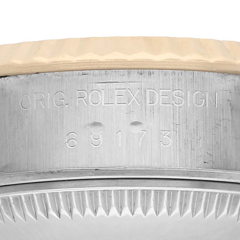 The image shows a close-up angle of the side of a Rolex Datejust 69173 Women
s Steel and Gold (two tone) Champagne Dial model, featuring engraved text and bezel detail.