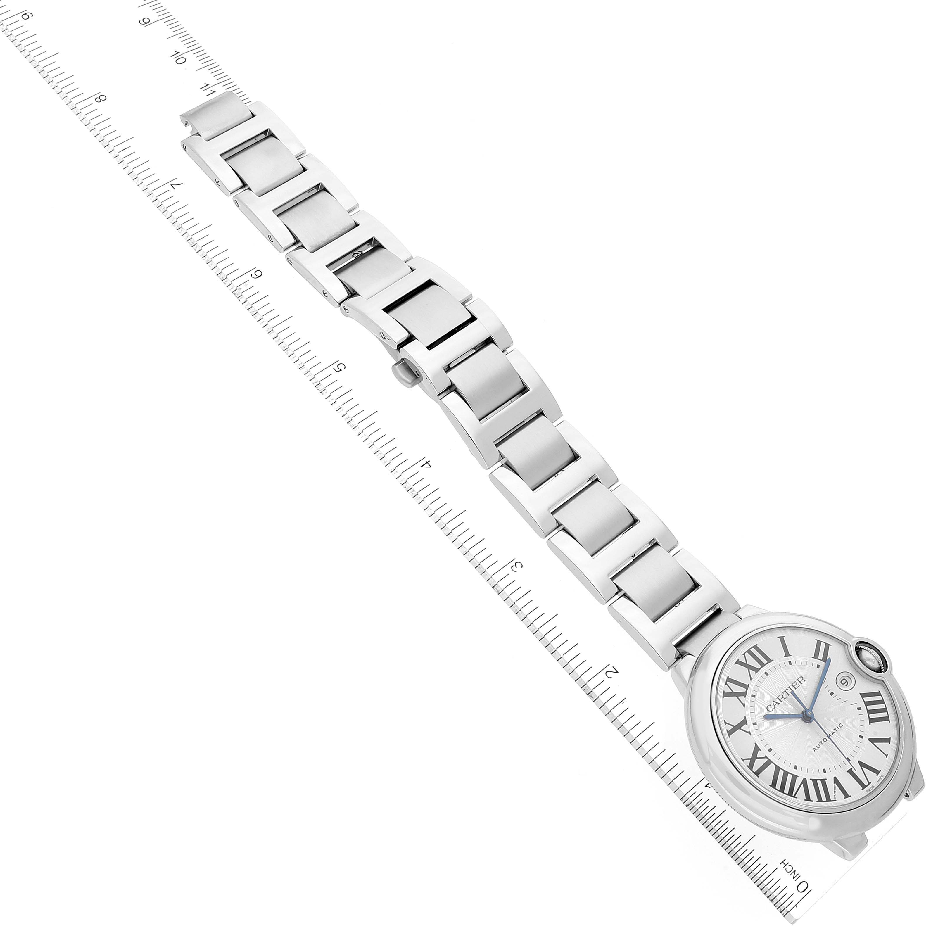 The image shows a Ballon Bleu watch by Cartier lying at an angle against a ruler, highlighting its face and full metal bracelet.
