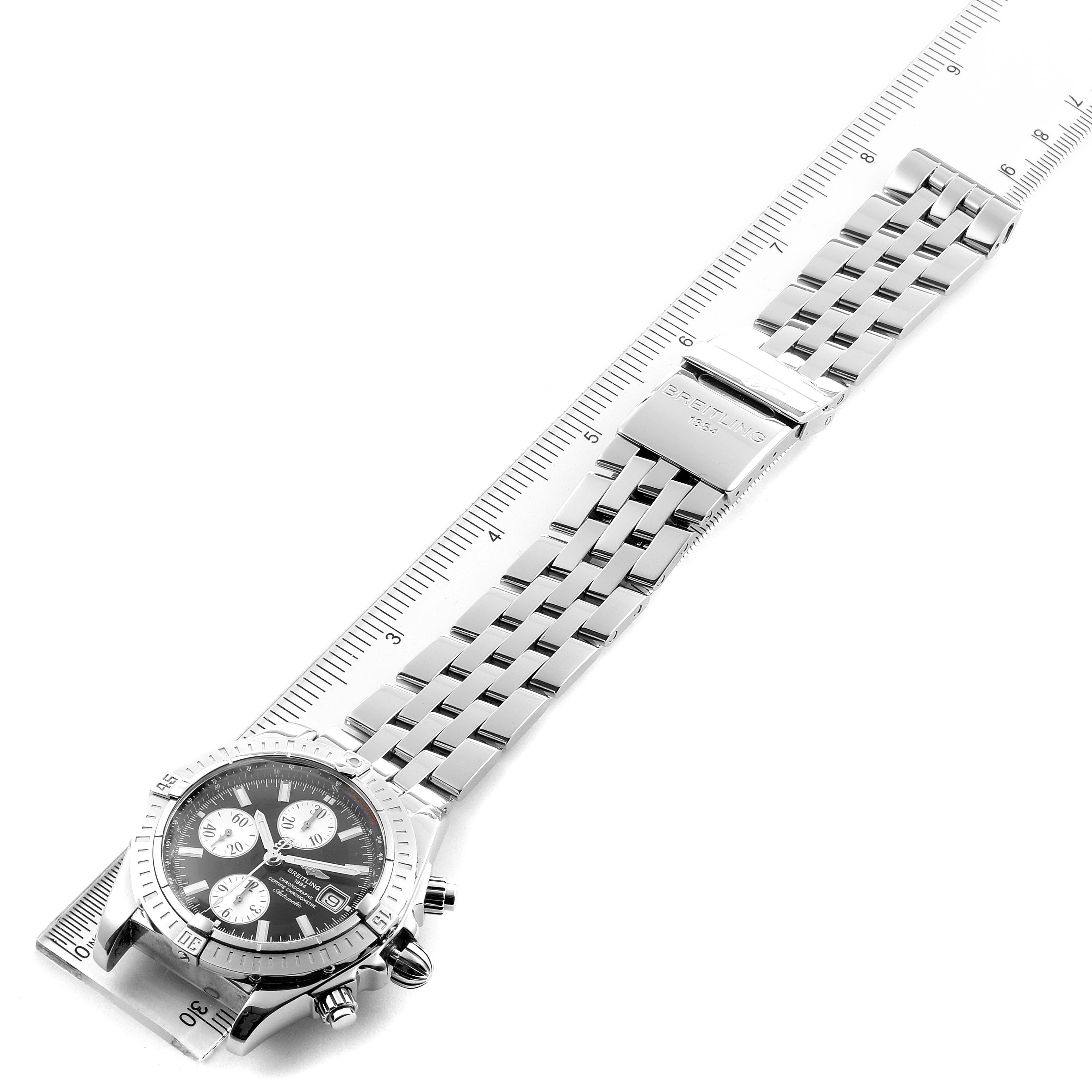The Breitling Chronomat A13356 Men
s Stainless Steel Black Dial A13356 Men
s Stainless Steel Black Dial watch is shown from a top-down angle, highlighting its face and metal bracelet extended alongside a ruler.
