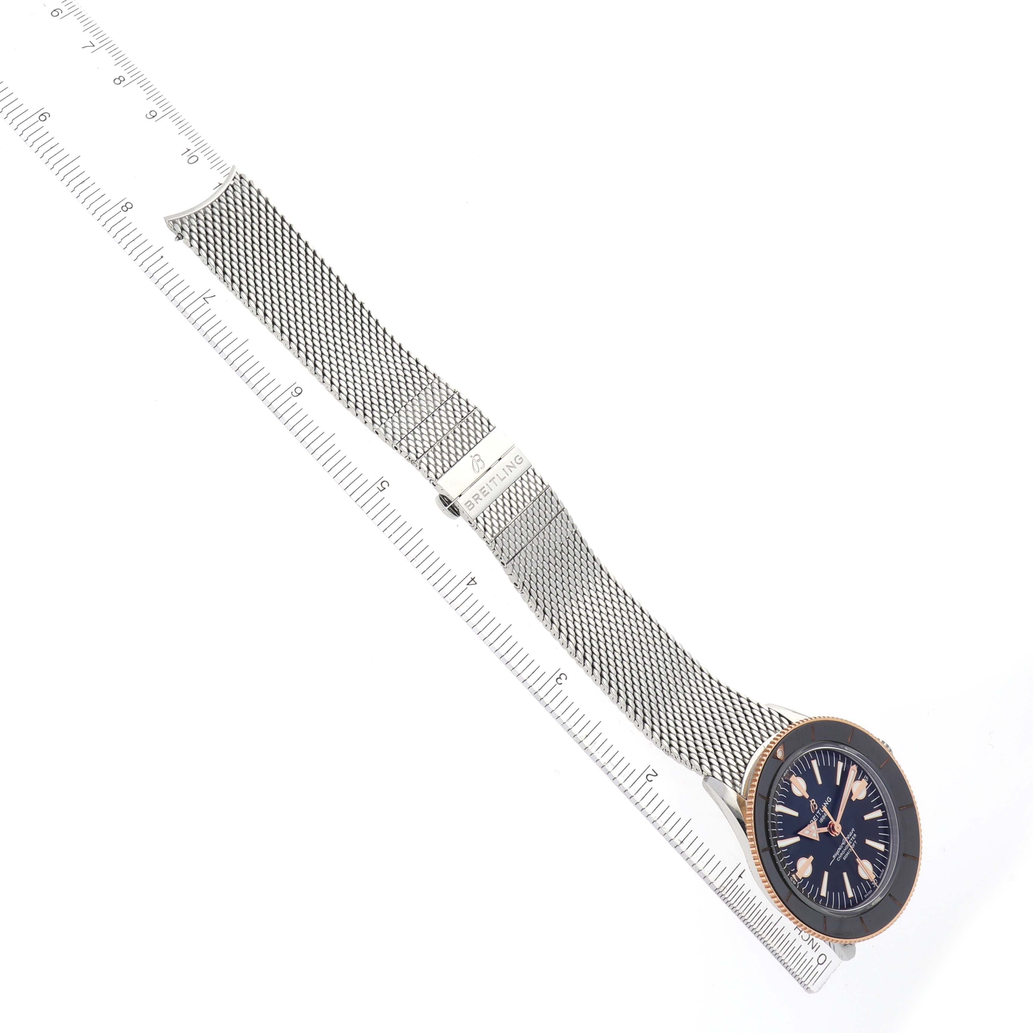 The Breitling Superocean U10370121B1A1 Men's Stainless Steel Black Dial watch is displayed from a top angle, showing its face, metal mesh strap, and clasp alongside a ruler.