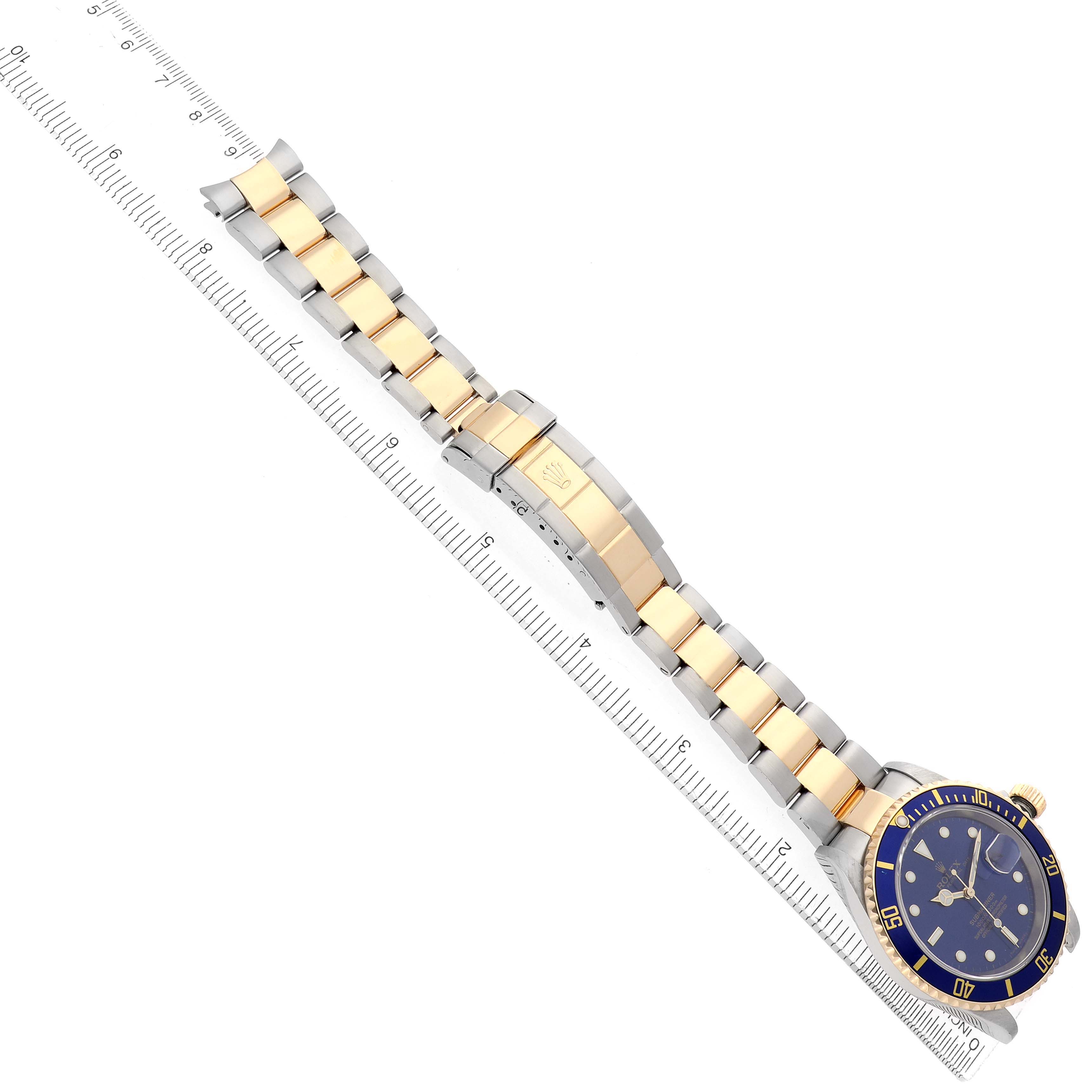 The Rolex Submariner 16613 Men's Steel and Gold (two tone) Blue Dial is shown from an overhead angle, displaying the blue dial, gold and silver bracelet, and clasp beside a ruler.