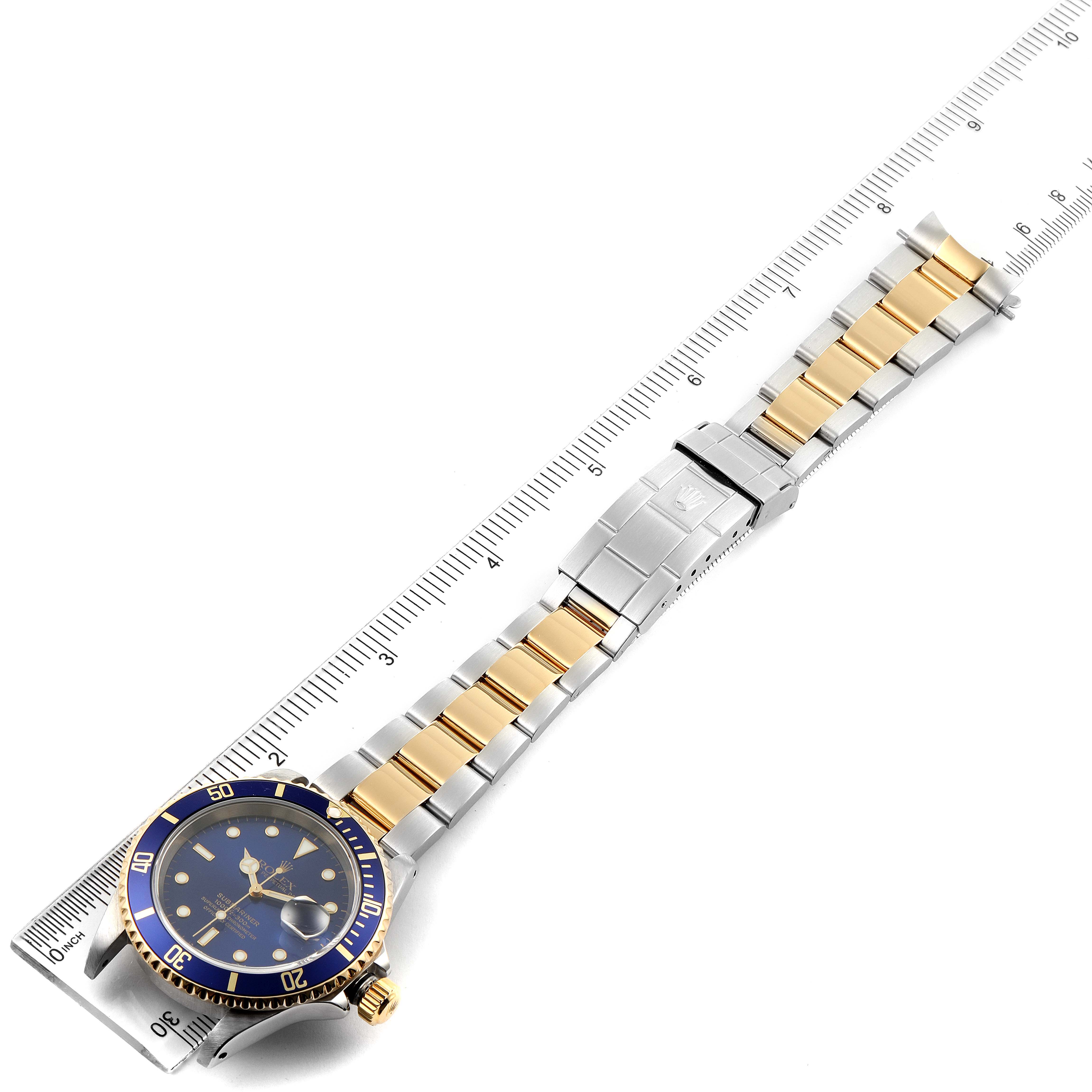 The image shows a Rolex Submariner 16613 Men's Steel and Gold (two tone) Blue Dial 16613 Men's Steel and Gold (two tone) Blue Dial watch laid flat with a ruler underneath, displaying the face, bracelet, and clasp.