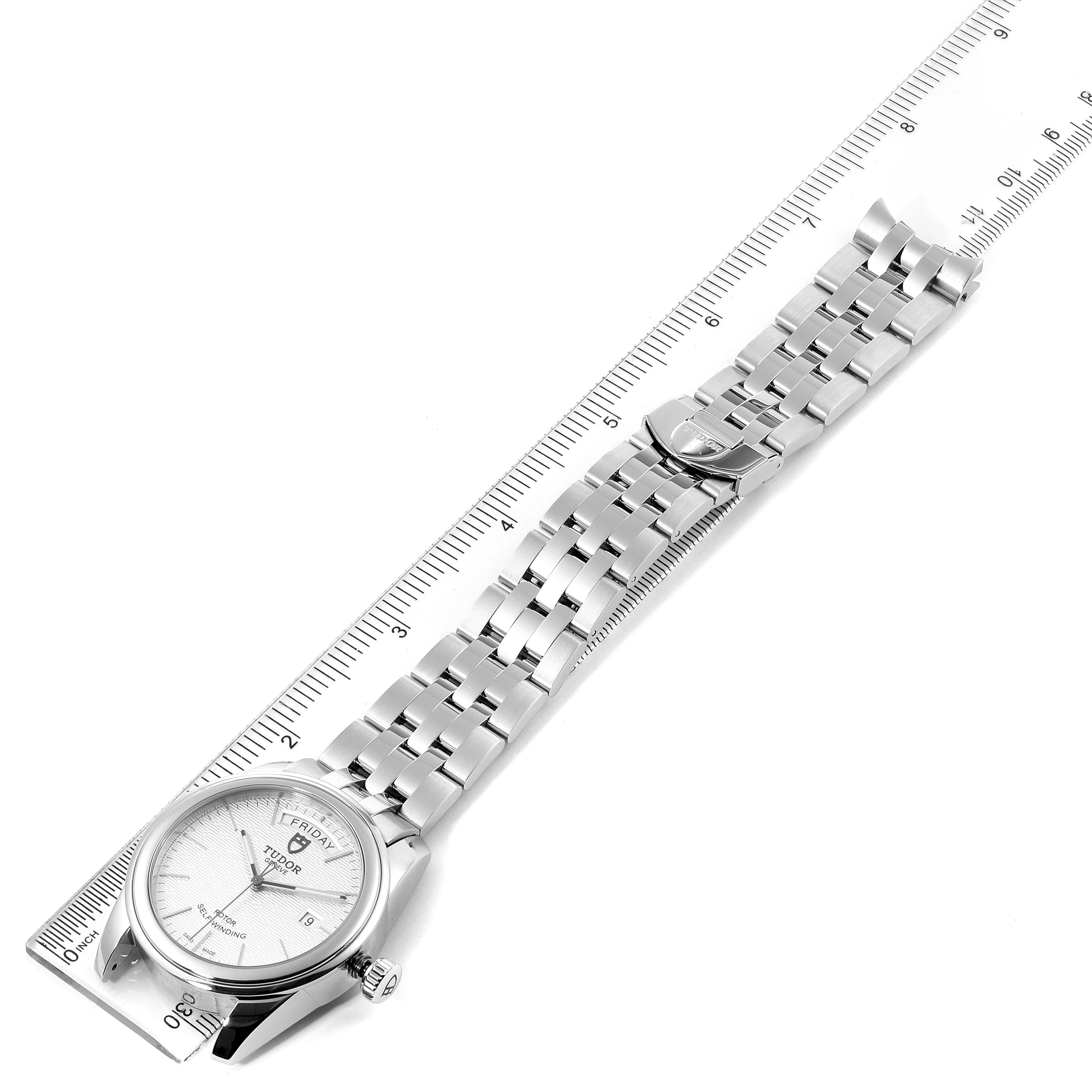 The image shows a top-down view of the Tudor Glamour M56000-0008 Men's Stainless Steel Silver Dial M56000-0008 Men's Stainless Steel Silver Dial model watch, including the face, bezel, bracelet, and clasp against a ruler.