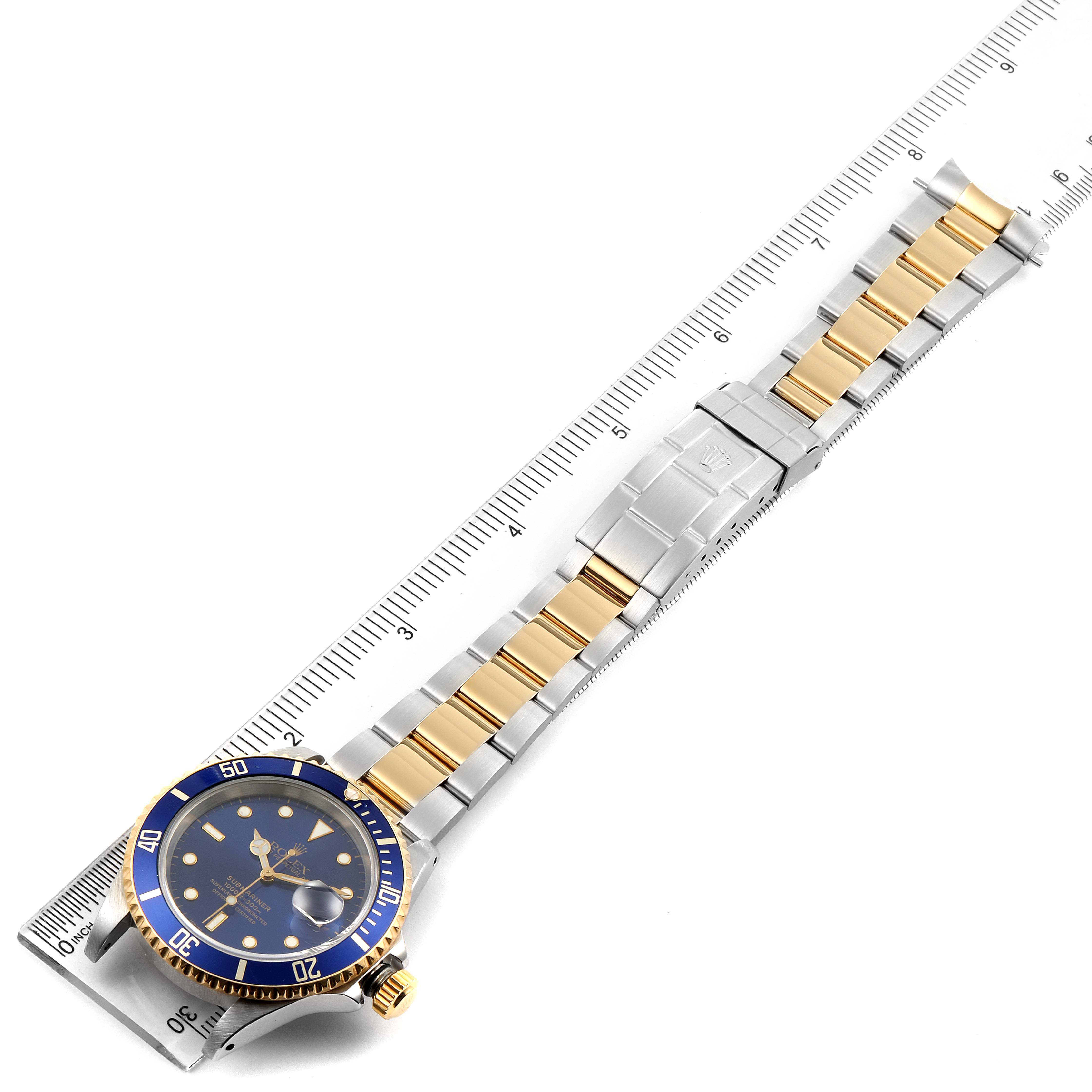 The image shows a Rolex Submariner 16613 Men's Steel and Gold (two tone) Blue Dial 16613 Men's Steel and Gold (two tone) Blue Dial watch laid flat alongside a ruler, highlighting its blue bezel, dial, and two-tone bracelet.