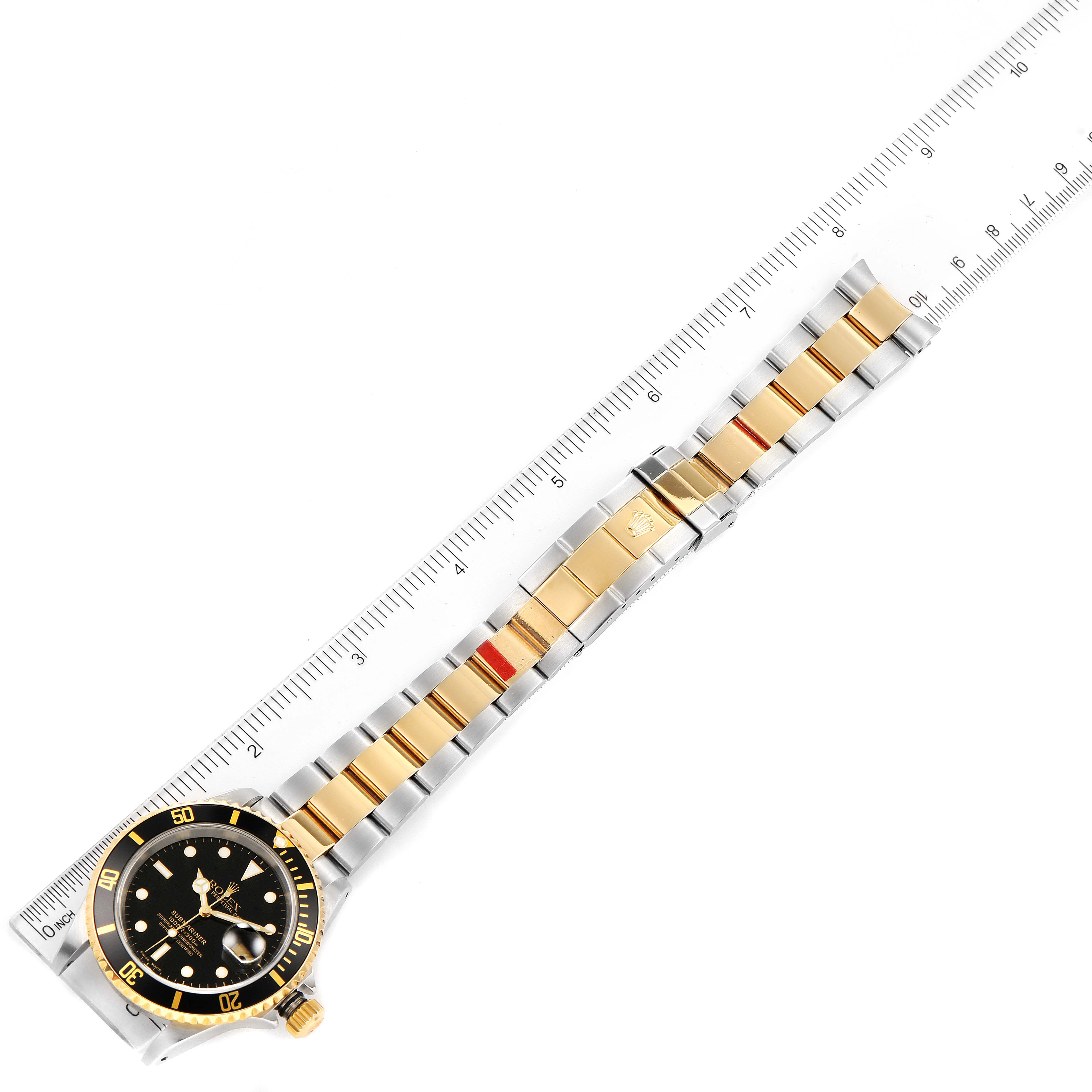 The image shows a Rolex Submariner 16613 Men's Steel and Gold (two tone) Black Dial 16613 Men's Steel and Gold (two tone) Black Dial watch laid flat alongside a ruler, highlighting its bracelet and face.