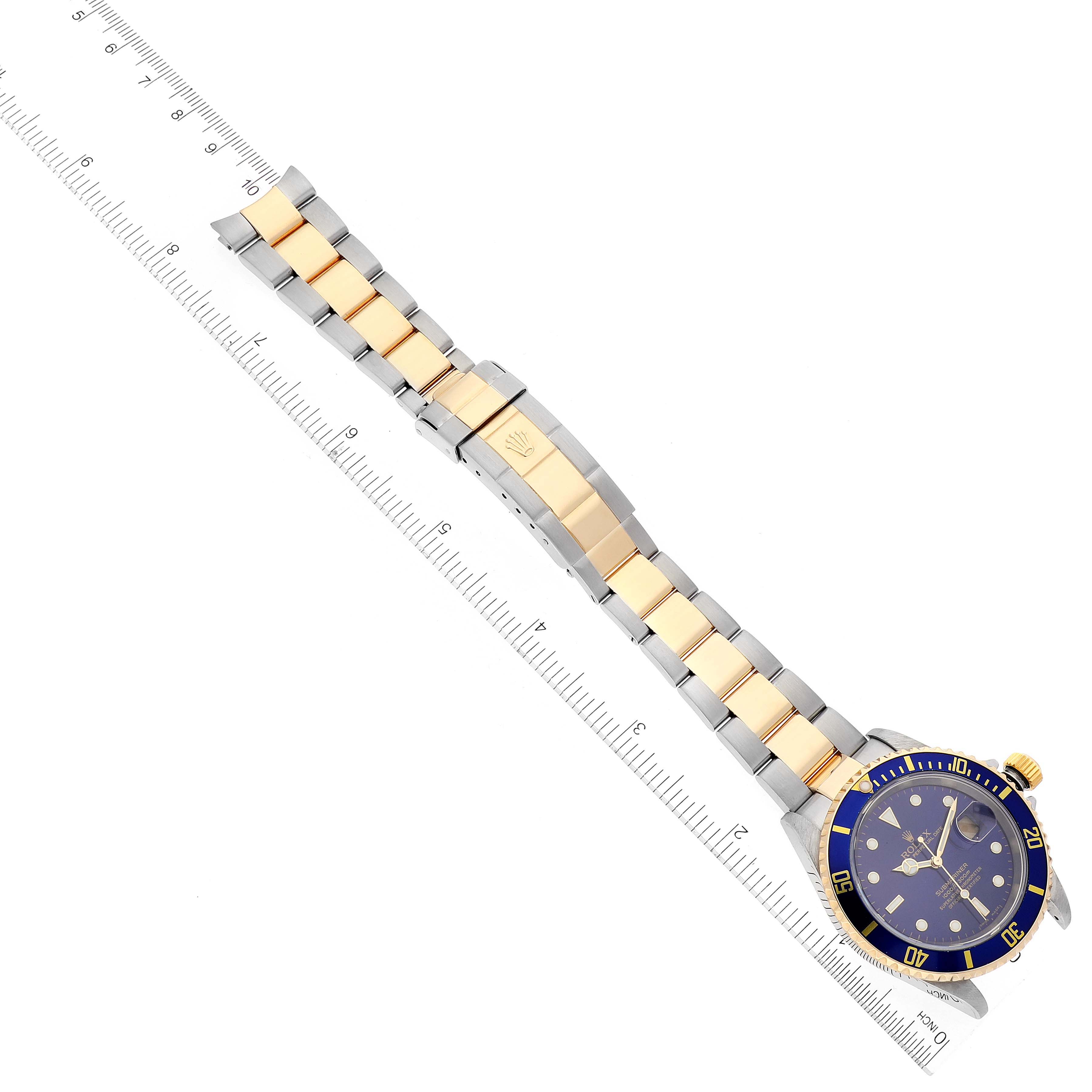 The image shows an overhead view of a Rolex Submariner 16613 Men's Steel and Gold (two tone) Blue Dial 16613 Men's Steel and Gold (two tone) Blue Dial watch with a blue dial and a two-tone band alongside a ruler for scale.