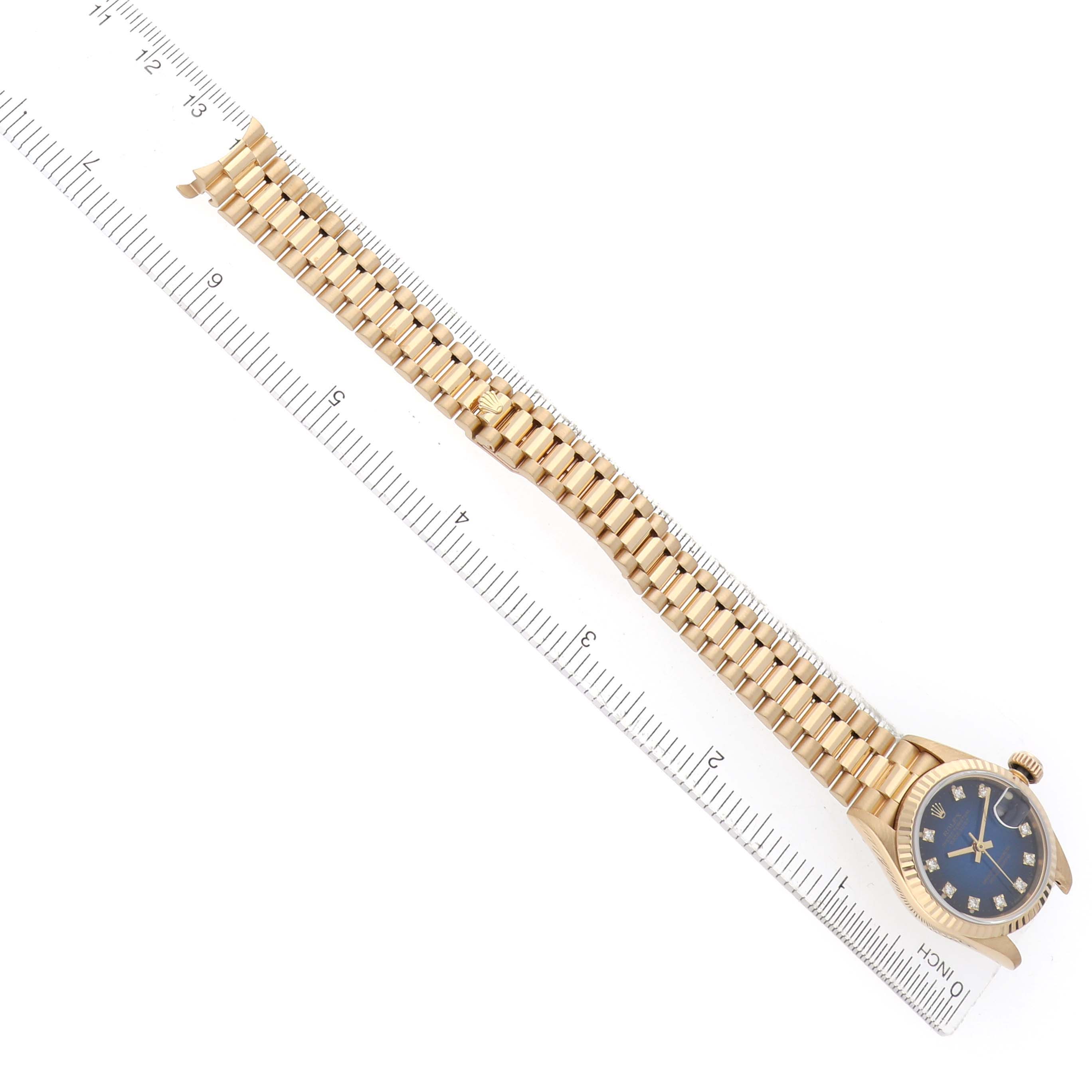The image shows a Rolex President 69178 Women's Yellow Gold Blue Dial model watch laid flat with its band extended alongside a ruler.