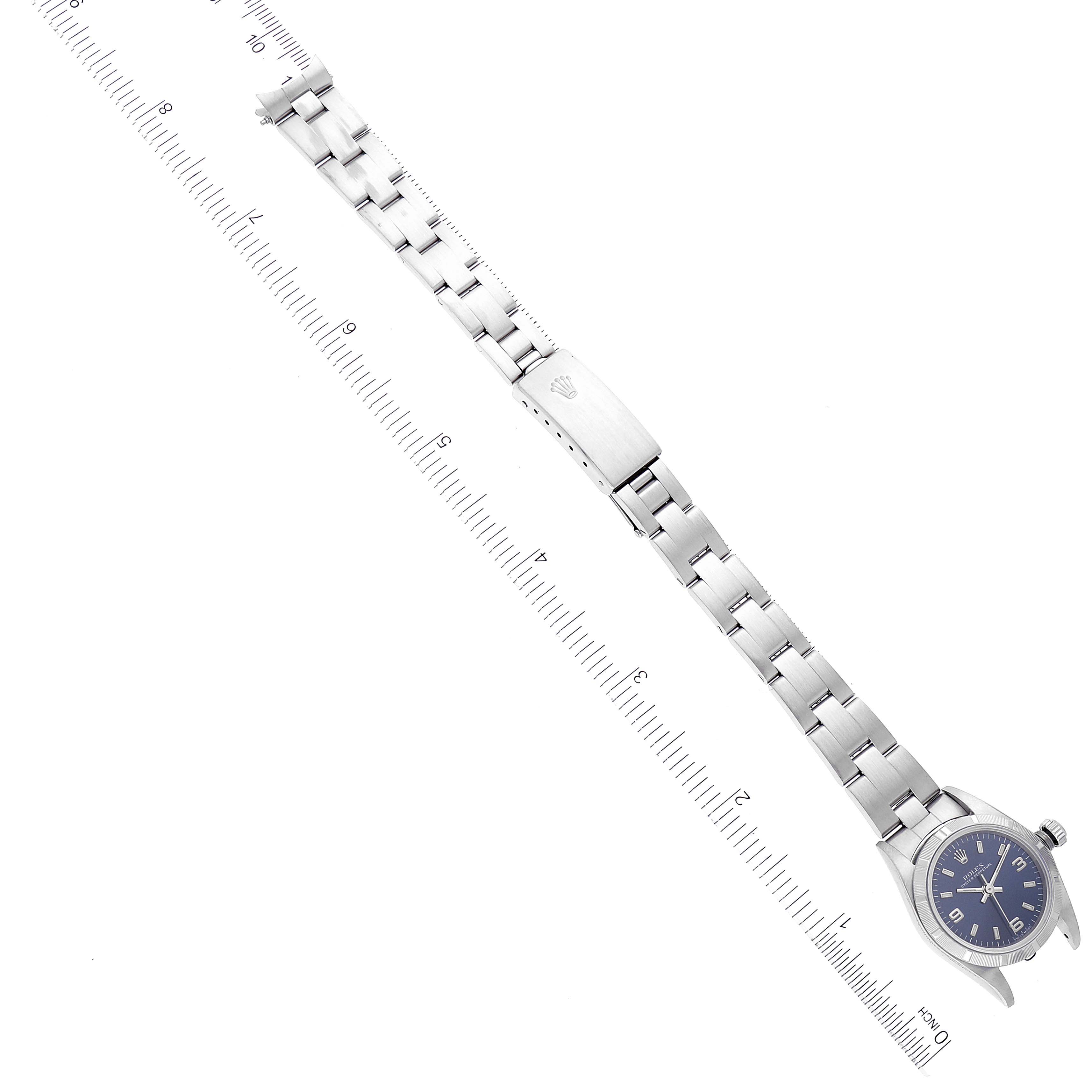 The Oyster Perpetual Rolex watch is shown from an angled top-down view, highlighting the face, bracelet, and clasp against a measuring scale.