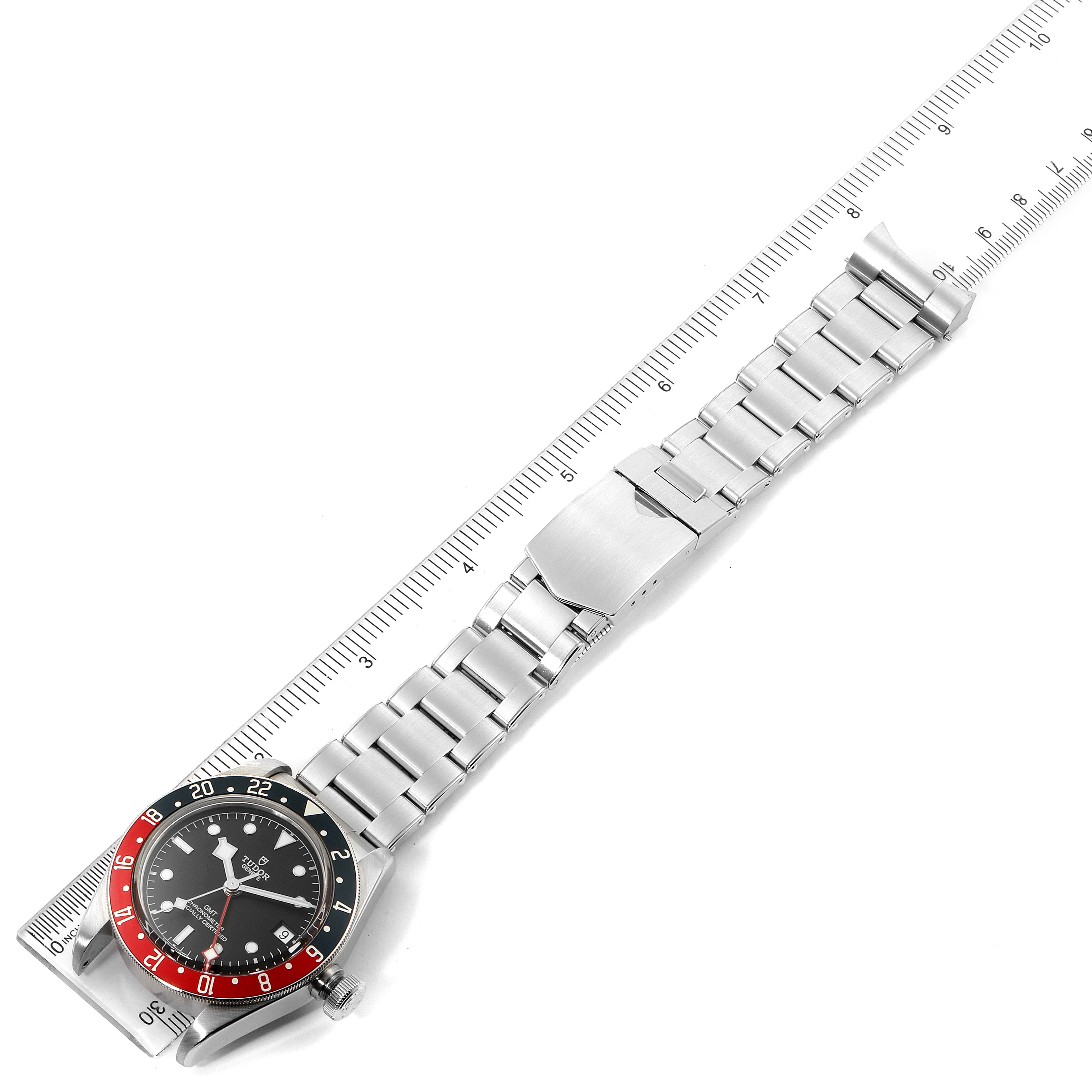 The image shows a Tudor Heritage Black Bay 79830RB Men
s Stainless Steel Black Dial 79830RB Men
s Stainless Steel Black Dial watch with a red and blue bezel and a stainless steel bracelet, placed beside a ruler.