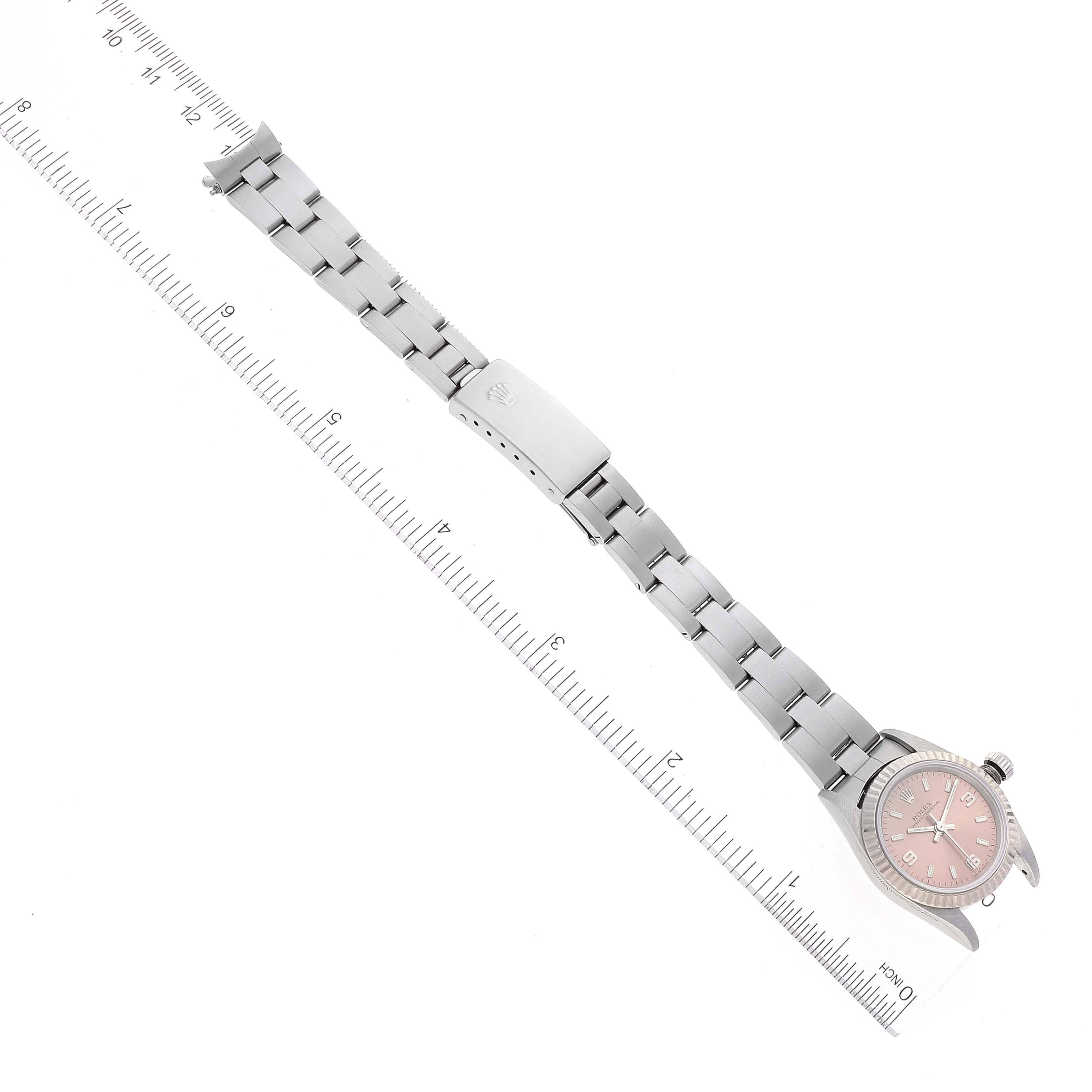 The Rolex Oyster Perpetual 76094 Women
s Stainless Steel Pink Dial 76094 Women
s Stainless Steel Pink Dial watch is shown from a top-down angle, with the band fully extended and positioned beside a ruler.