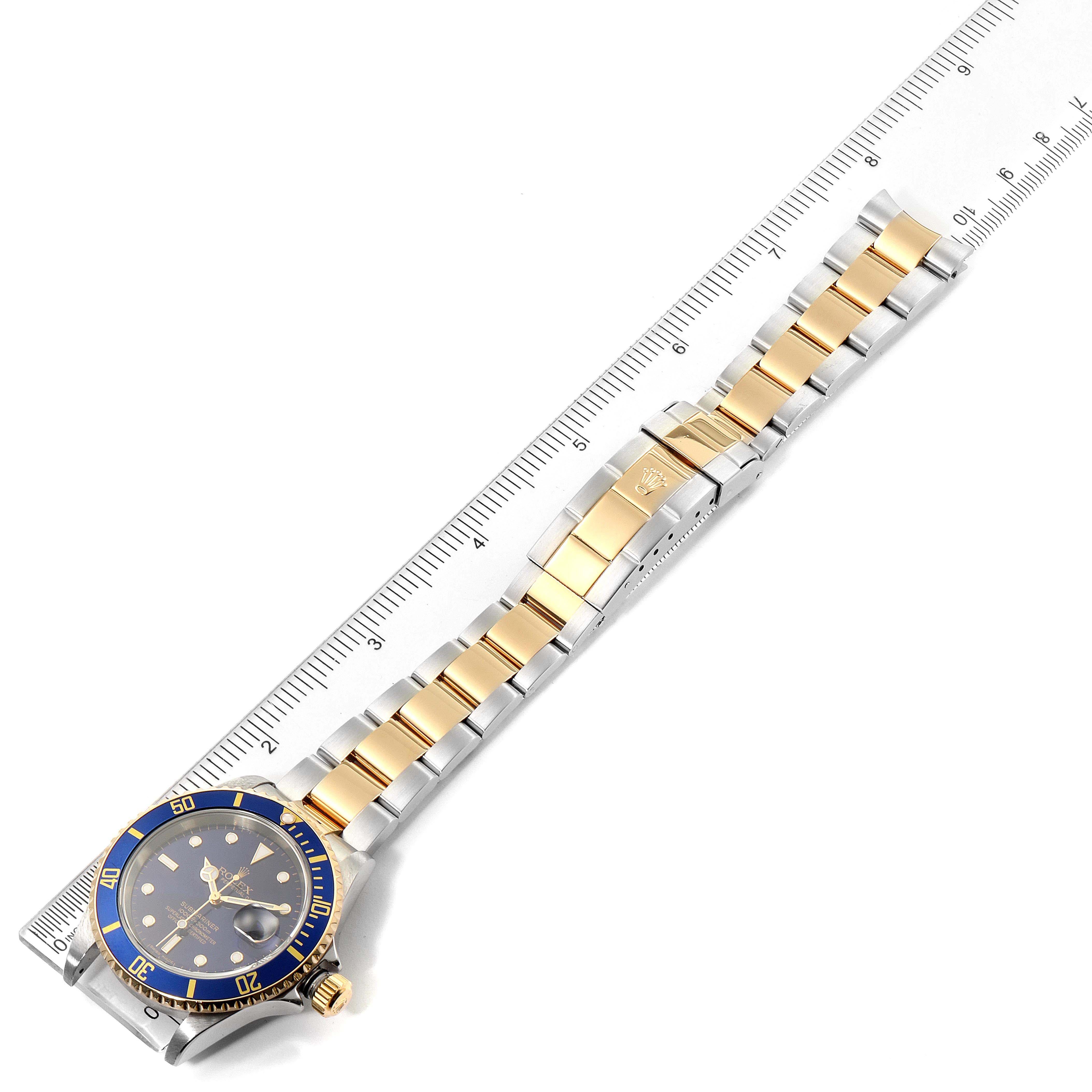 The image shows a top-down view of a Rolex Submariner 16613 Men's Steel and Gold (two tone) Blue Dial 16613 Men's Steel and Gold (two tone) Blue Dial watch, including its face and two-tone metal bracelet next to a ruler.