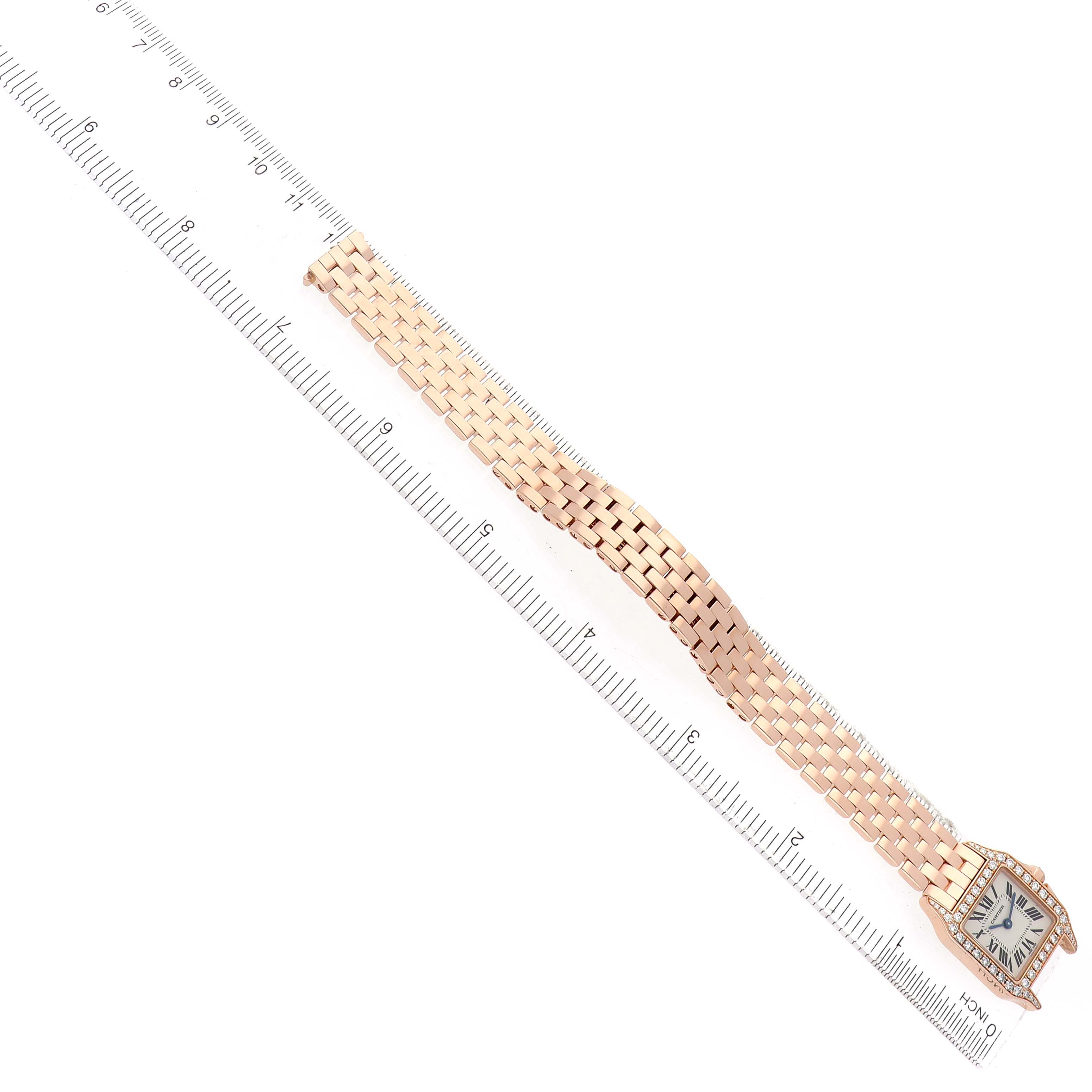 The Cartier Santos Demoiselle WF9011Z8 Women's Rose Gold Silver Dial watch is shown from a top-down angle, displaying its face and gold bracelet alongside a ruler.