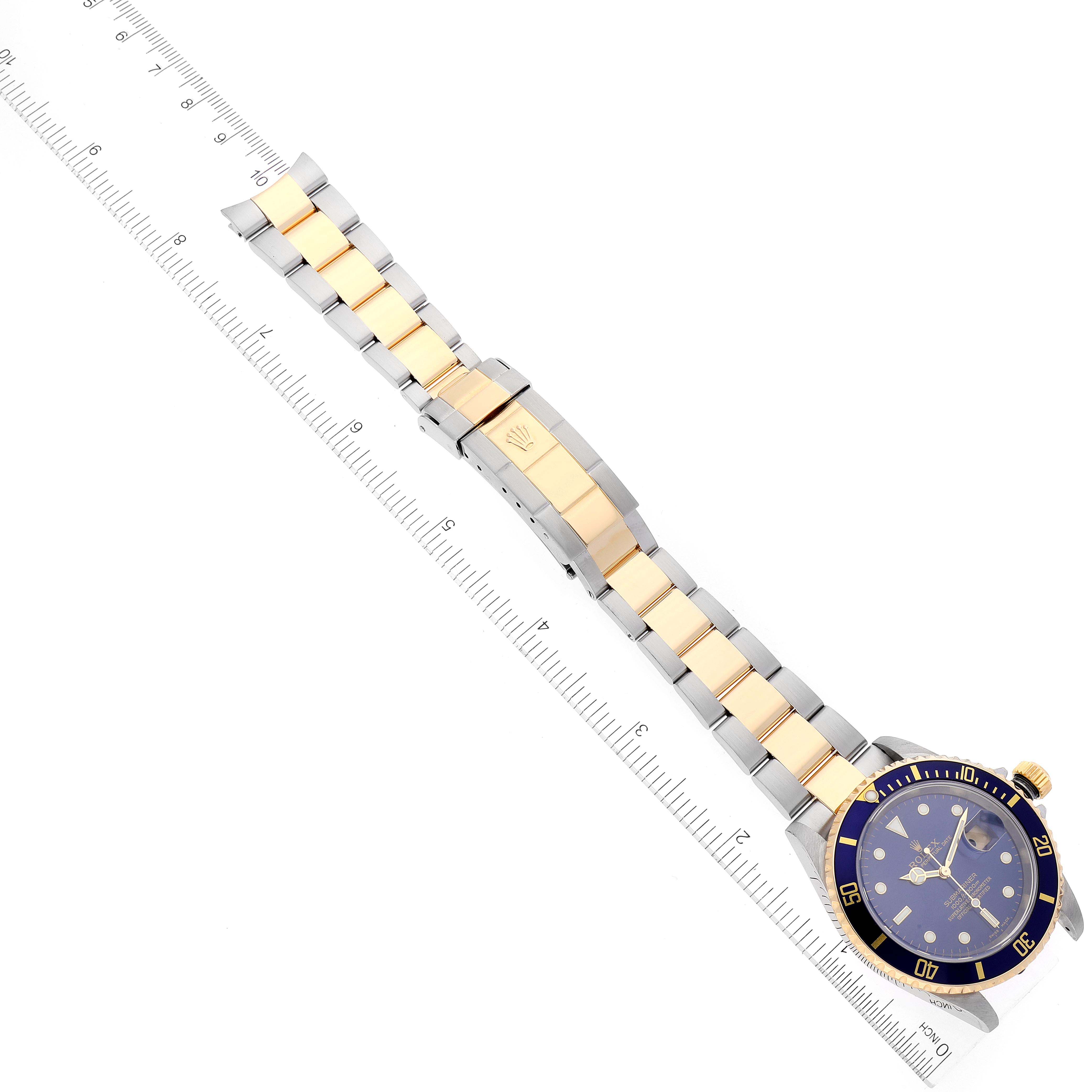 The image shows a Rolex Submariner 16613 Men's Steel and Gold (two tone) Blue Dial 16613 Men's Steel and Gold (two tone) Blue Dial watch laid flat alongside a ruler, displaying its bracelet, clasp, and dial face from a top-down angle.