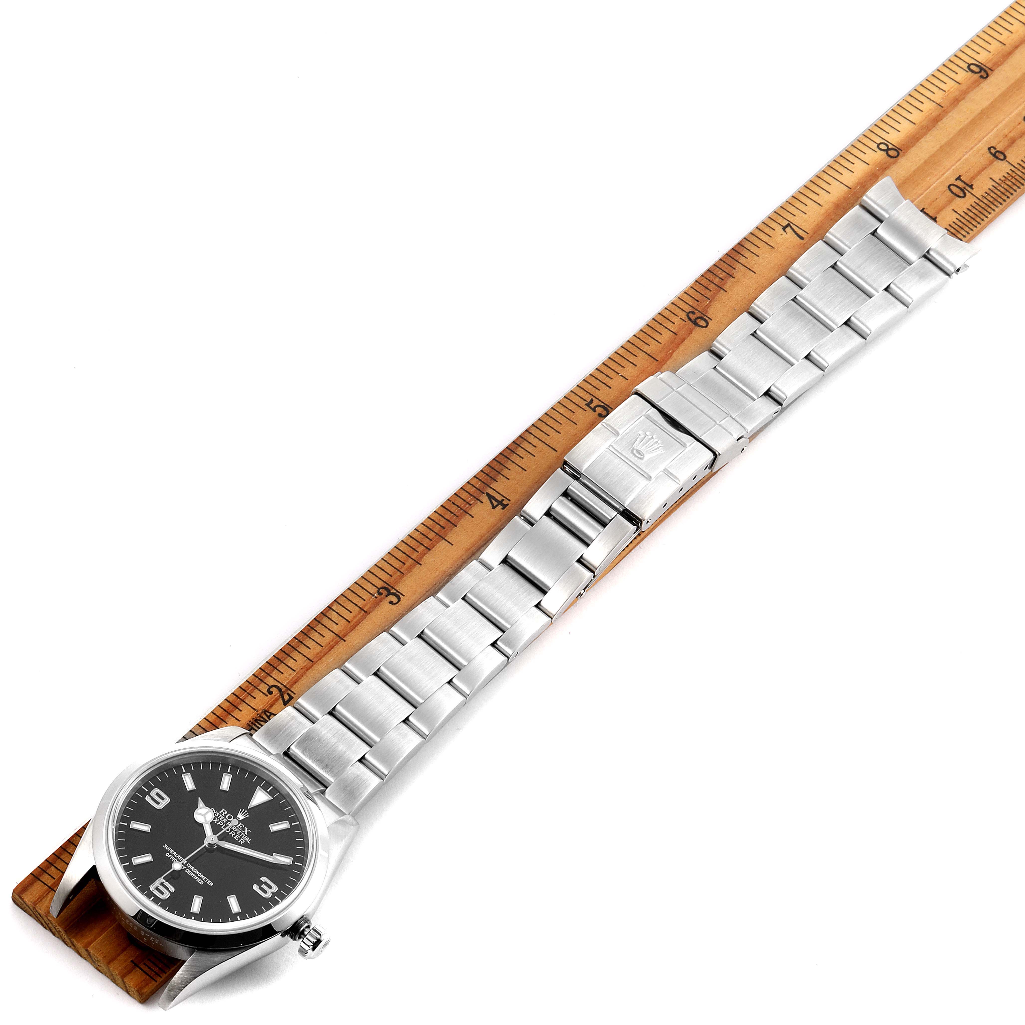 The Rolex Explorer 114270 Men
s Stainless Steel Black Dial 114270 Men
s Stainless Steel Black Dial watch is shown face-up, horizontally aligned with a ruler, displaying its dial, bracelet, and clasp.