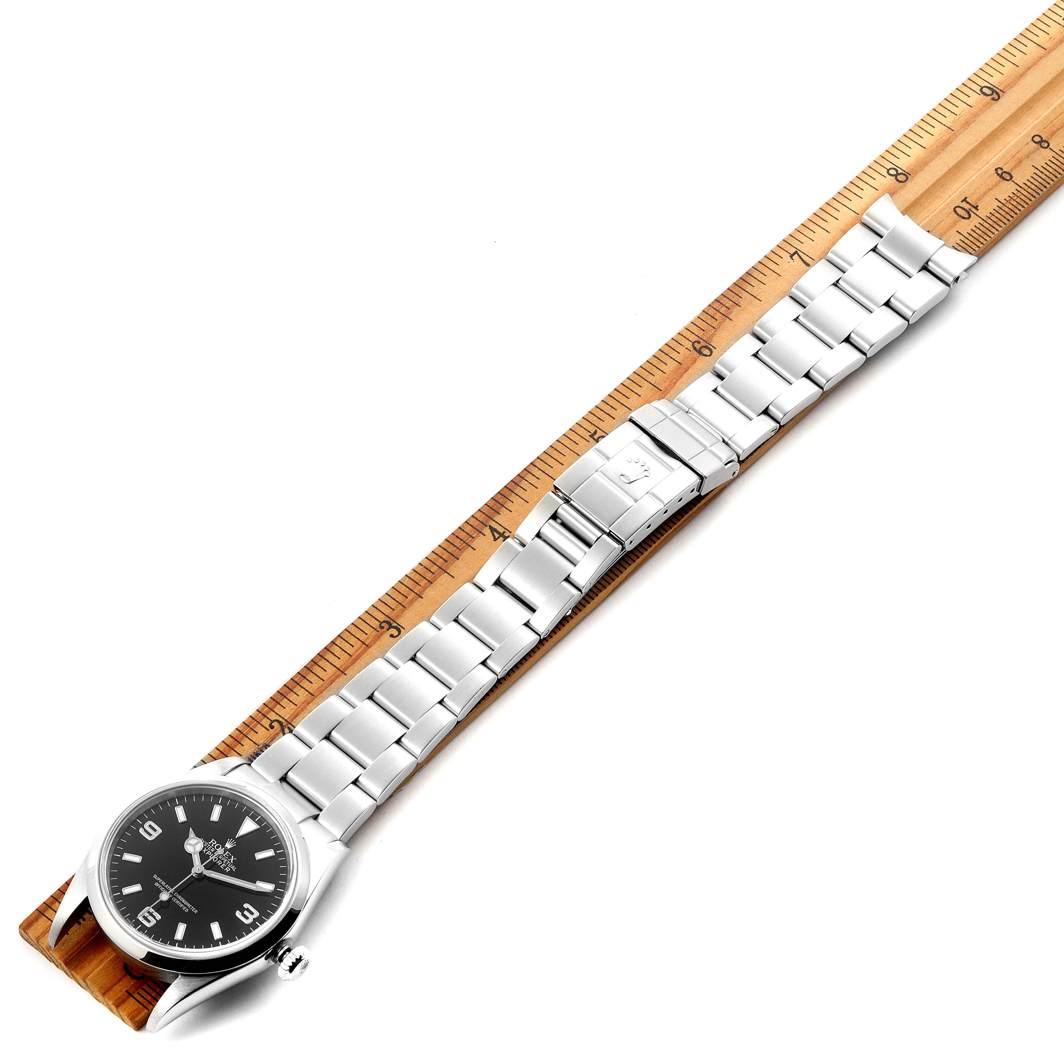The Rolex Explorer 114270 Men's Stainless Steel Black Dial 114270 Men's Stainless Steel Black Dial watch is shown at an overhead angle with its stainless steel bracelet and clasp laid out along a wooden ruler.