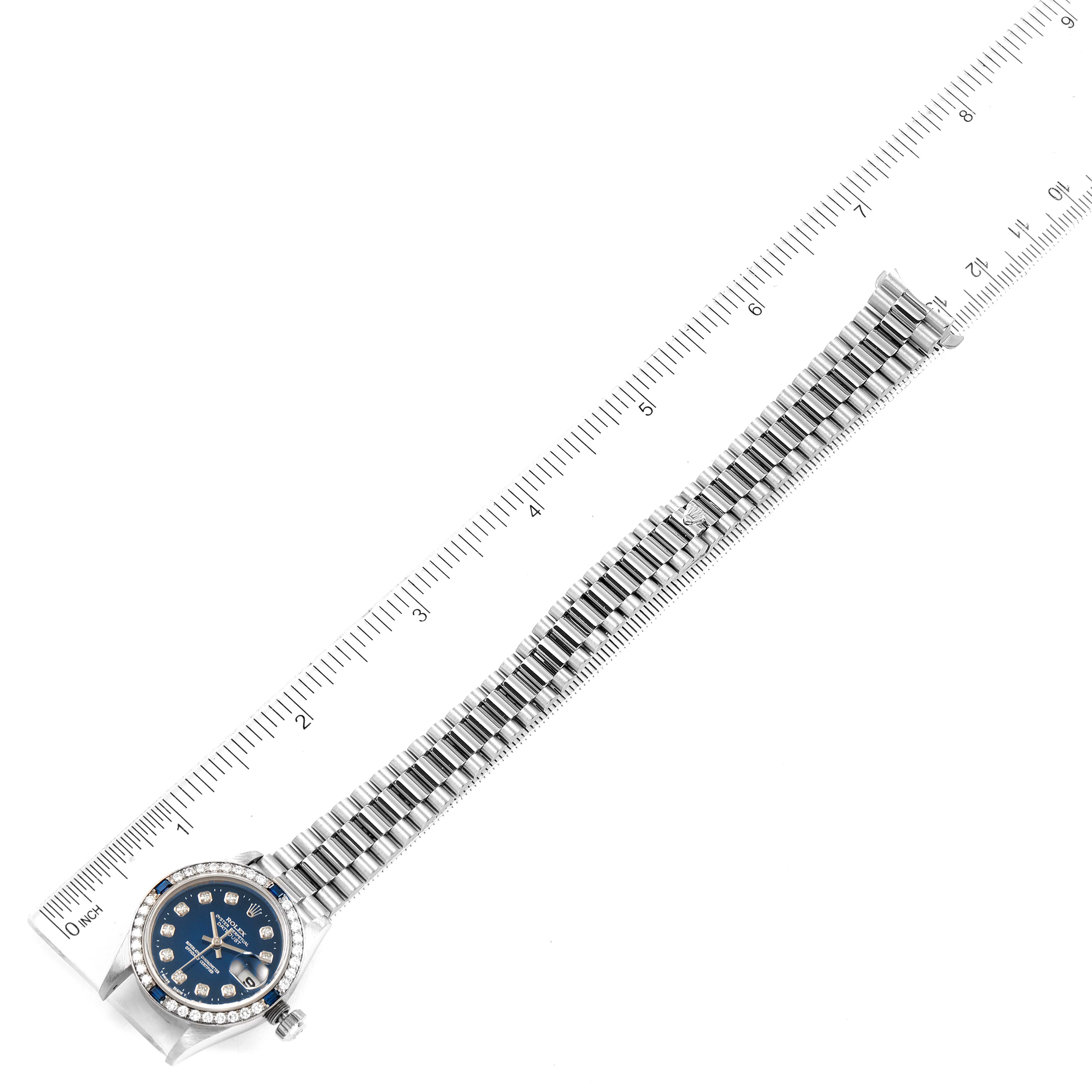 The image shows a top-down view of a Rolex President 79089 Women's White Gold Blue Dial 79089 Women's White Gold Blue Dial model watch, displaying the full strap and face next to a ruler for scale.