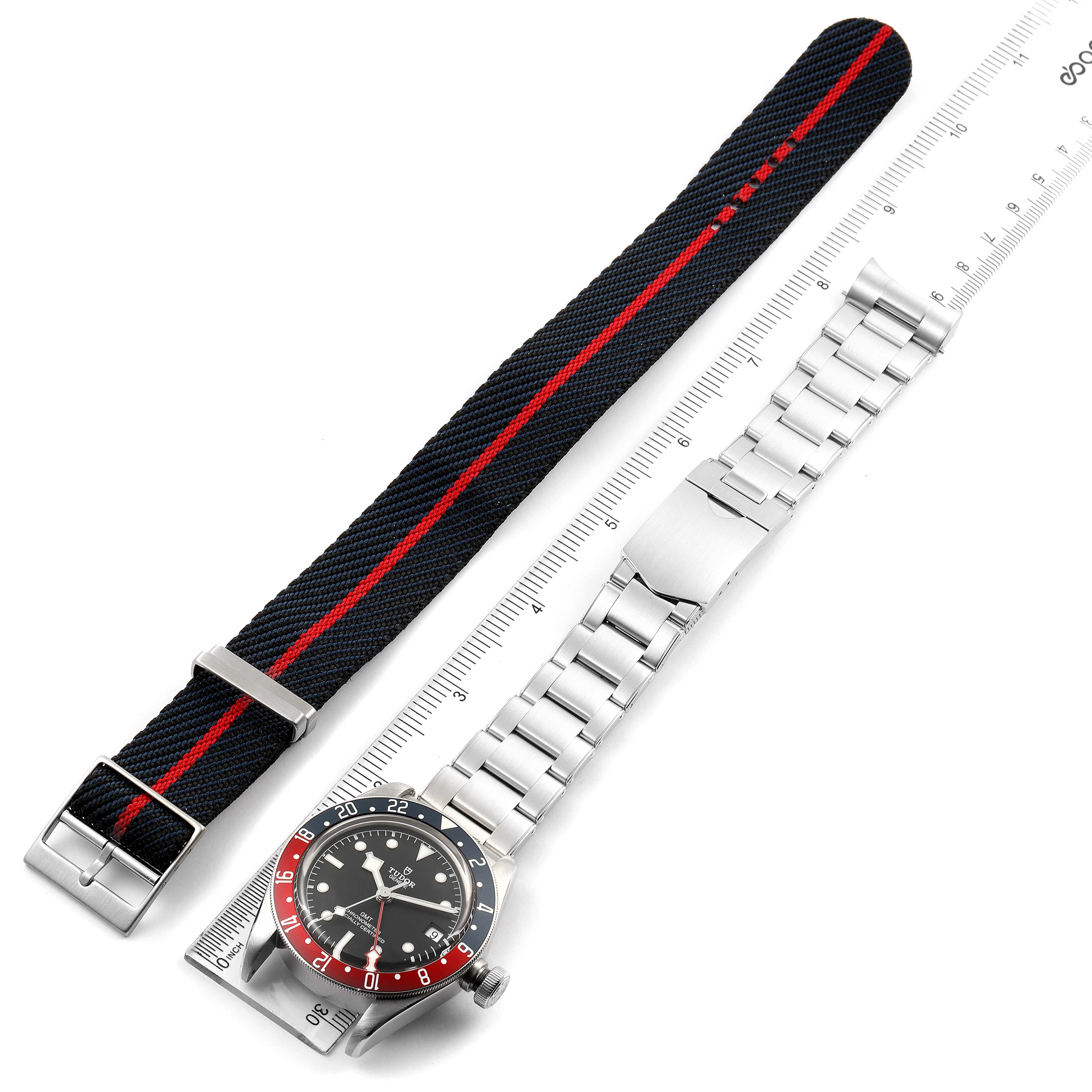 The image shows a Tudor Heritage Black Bay 79830RB Men
s Stainless Steel Black Dial 79830RB Men
s Stainless Steel Black Dial watch with a metal bracelet and an additional black fabric strap with a red stripe.