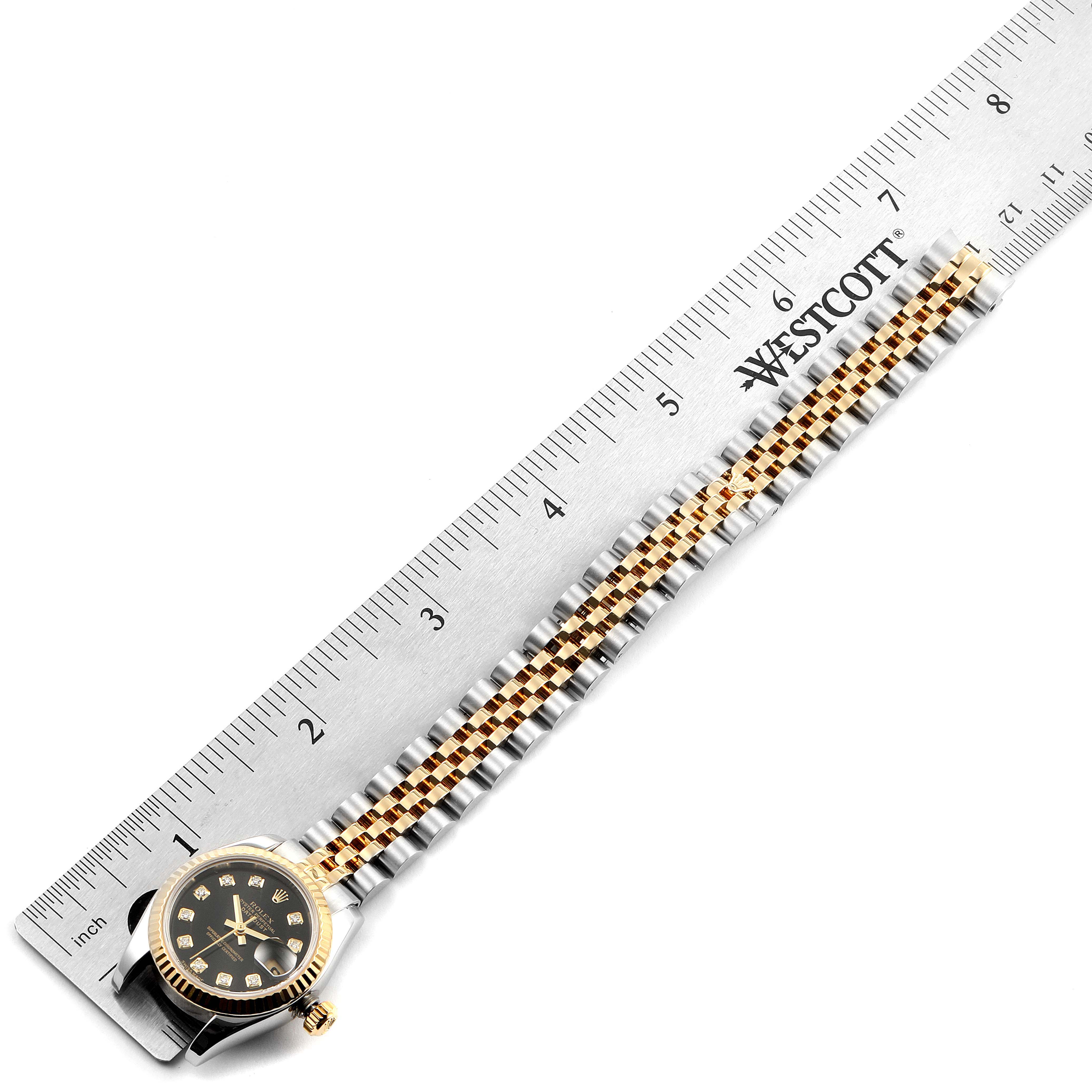The image shows a Rolex Datejust 179173 Women's Steel and Gold (two tone) Black Dial 179173 Women's Steel and Gold (two tone) Black Dial watch laid on a ruler, highlighting the watch face and band.